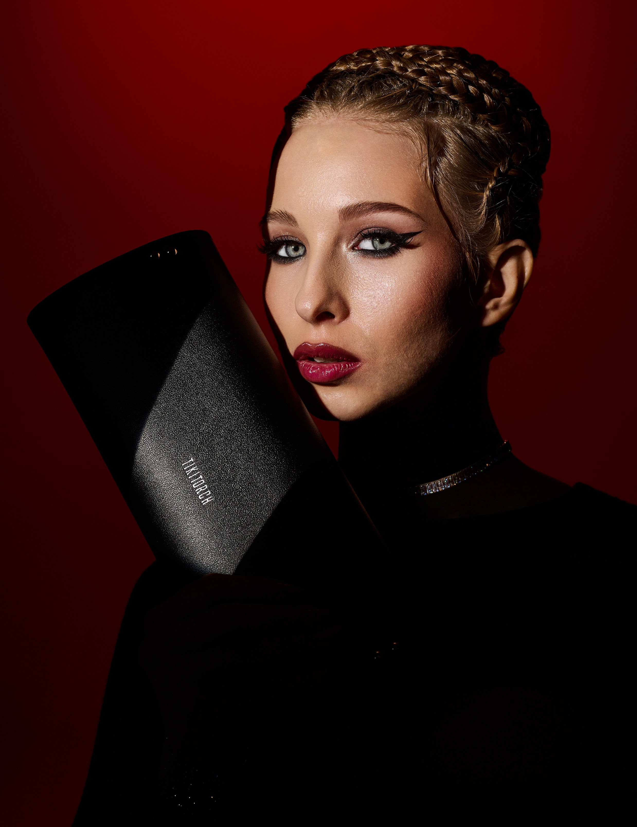 Портрет девушки с плетеными волосами, в темном наряде с ярким макияжем и черной сумкой-клатчем на фоне глубокого красного цвета