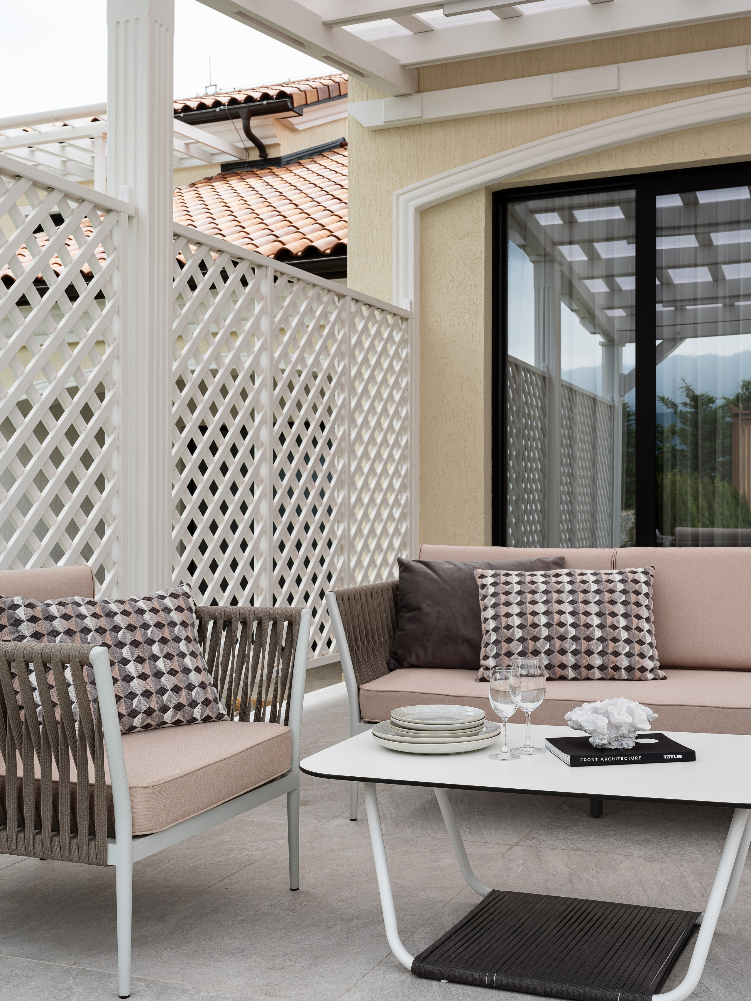 Уютная лаунж-зона на террасе с плетёной мебелью, геометрическими подушками и белым кофейным столиком, оформление фасада с белыми решётчатыми перегородками, автор проекта дизайнер Василисса Бачева, стилист Наталья Якушева, фото Сергей Денисенко.