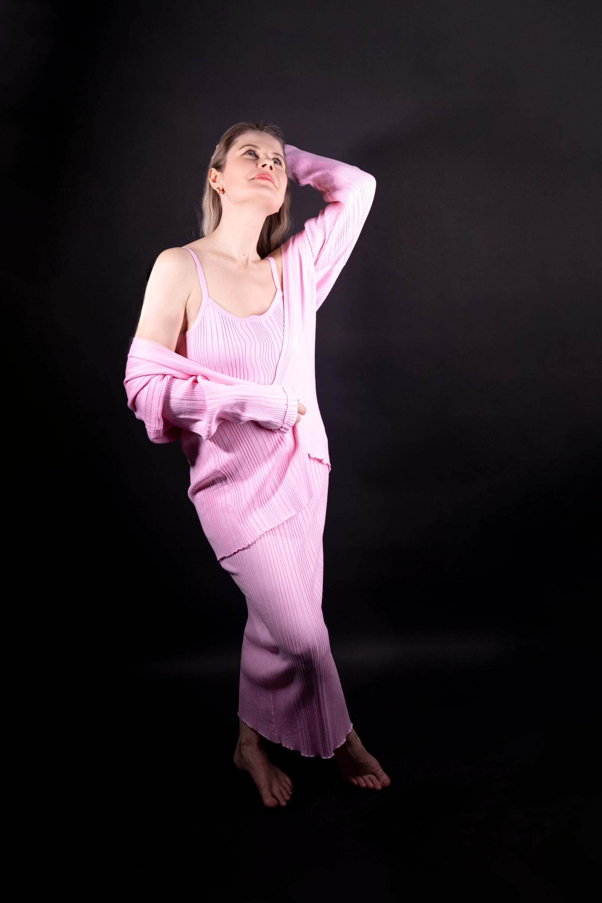 Момент лёгкости и свободы: женщина в нежном розовом трикотажном комплекте словно парит на тёмном фоне. Её поза полна грации и непринуждённости.