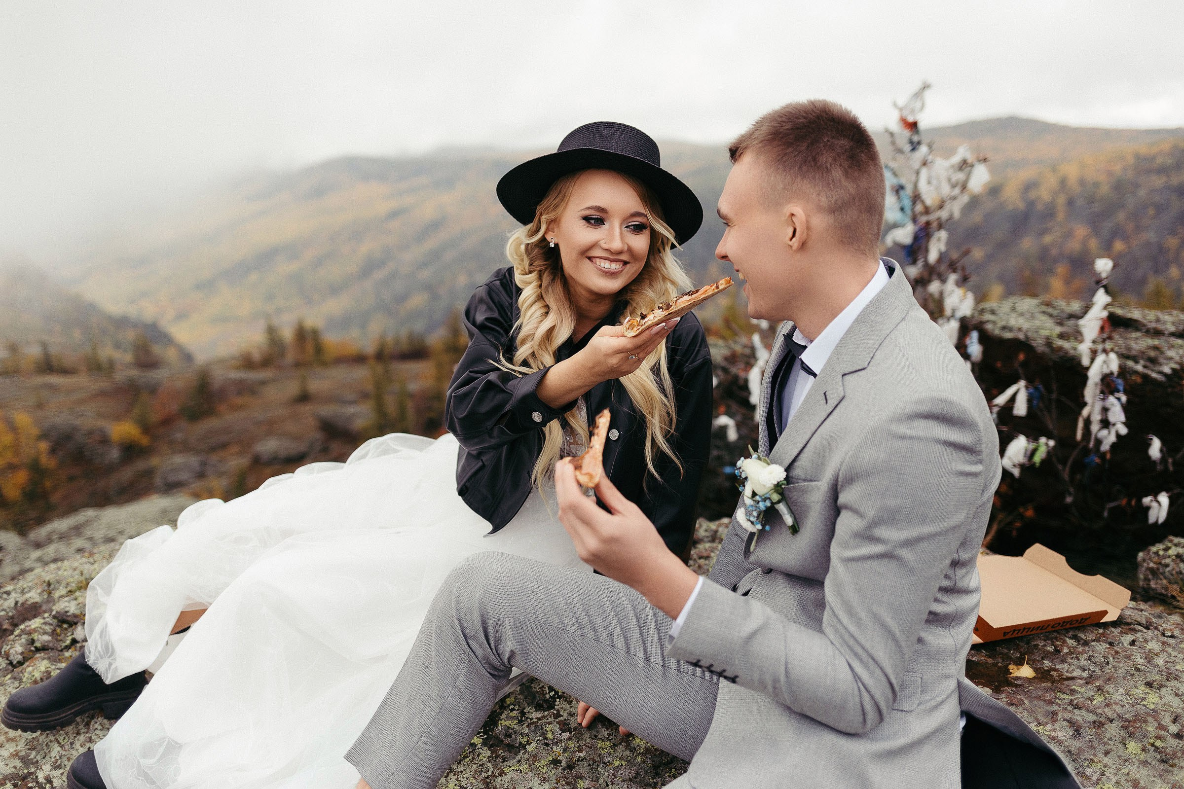 Невеста в шляпе кормит жениха пиццей сидя на скале на фоне гор. 