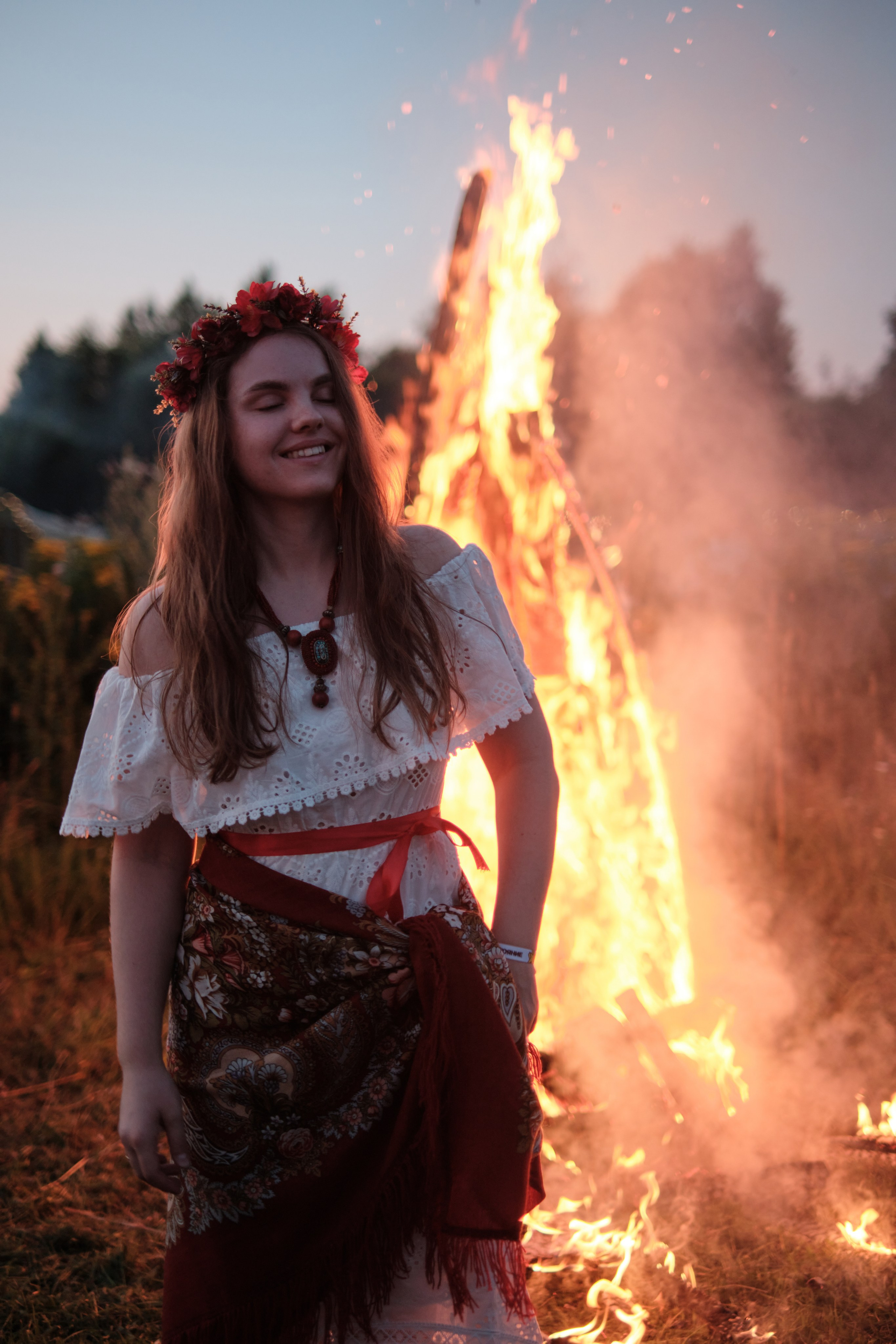 Девушка стоит в русском костюме и венком сзади горит большой костер