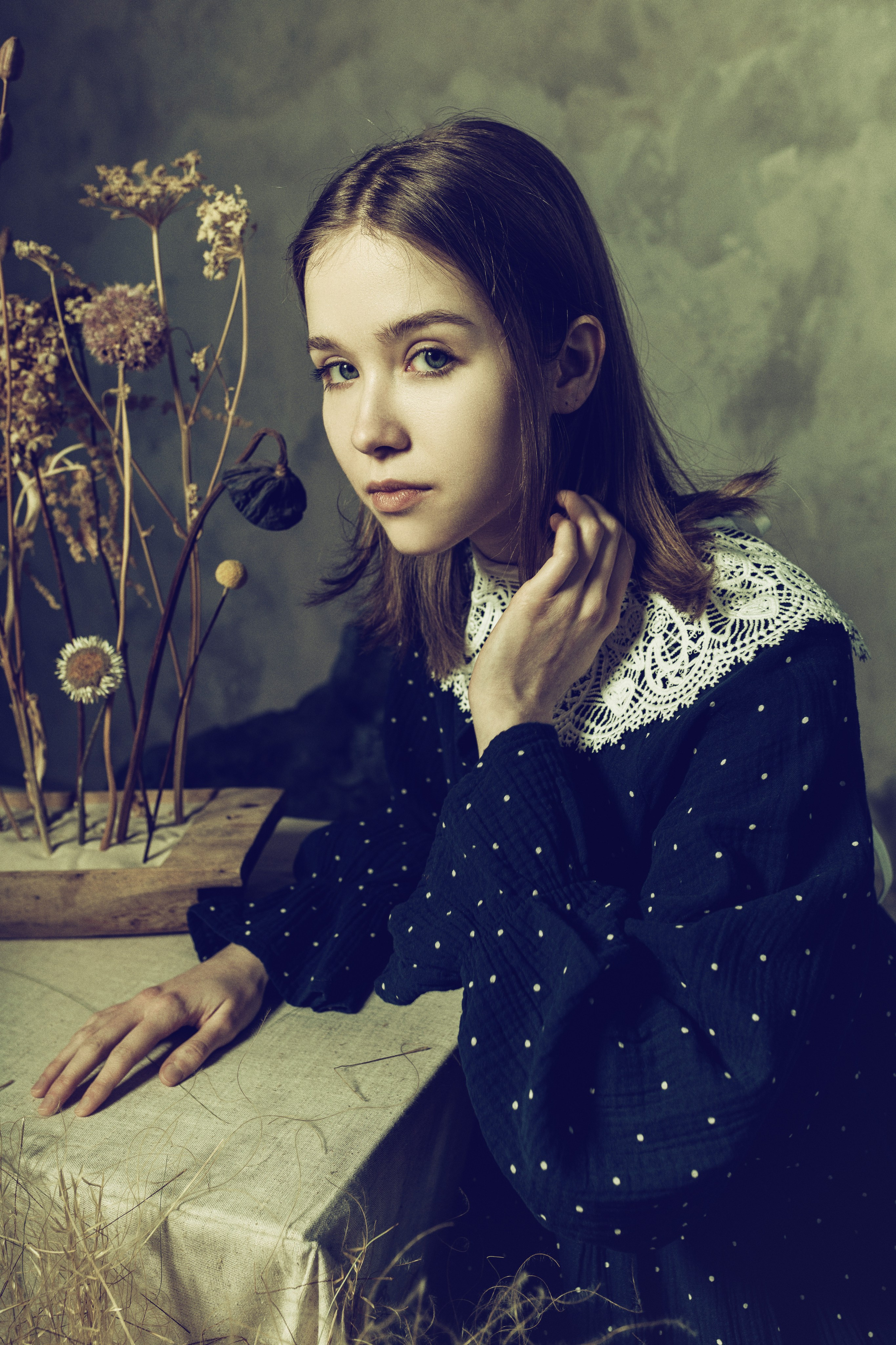 Молодая женщина в винтажном наряде с кружевным воротничком задумчиво сидит за столом, уставленным сухоцветами. Мягкое освещение создает созерцательное, ностальгическое настроение.
