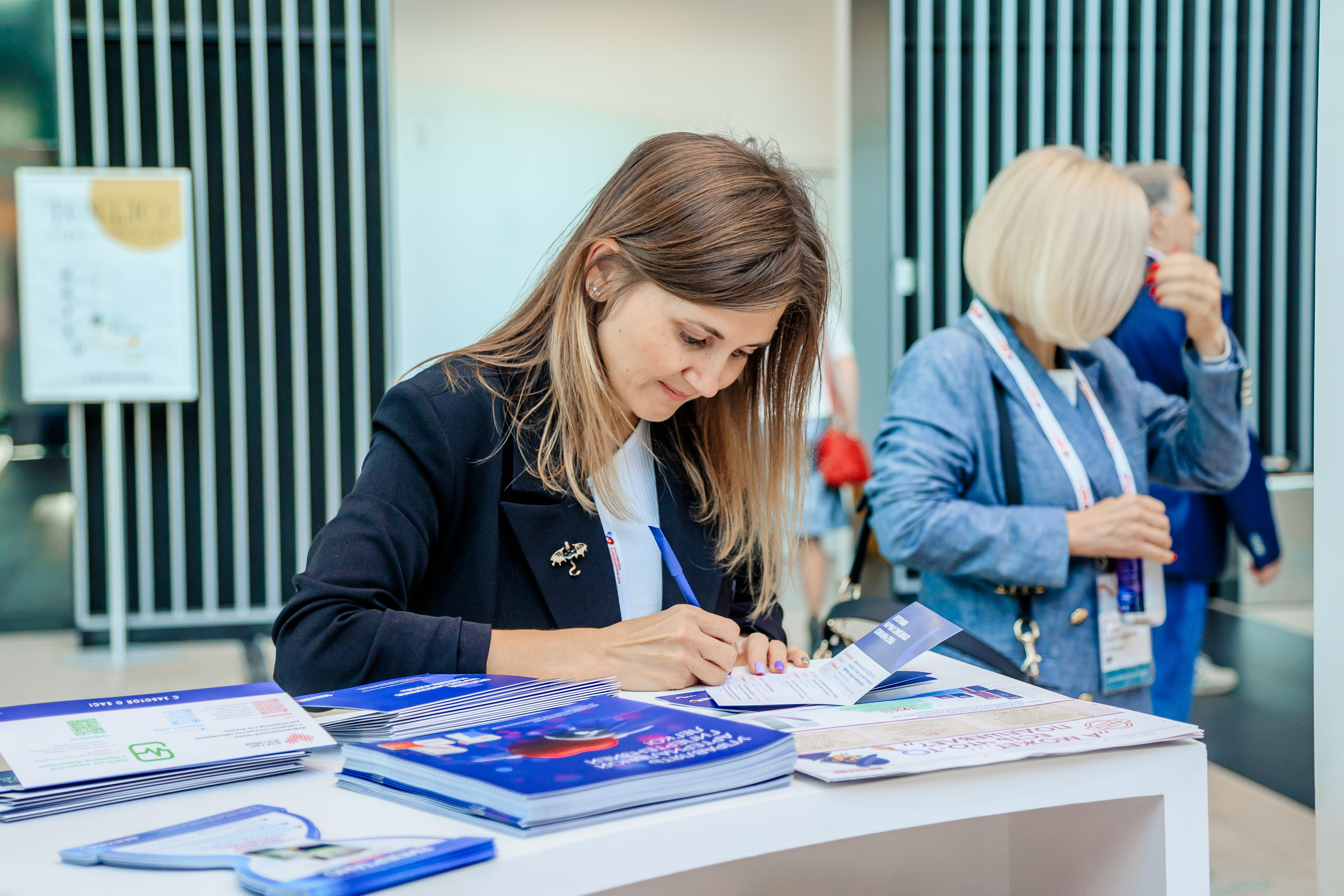 Женщина за информационной стойкой рекламного стенда делает запись в блокнот, сзади идут люди