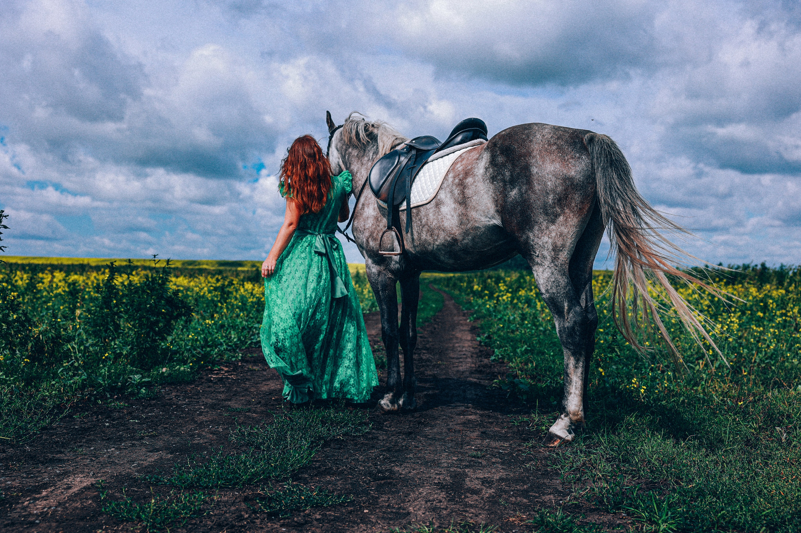 Женщина в длинном зеленом платье стоит рядом с седланной серой лошадью на проселочной дороге в поле с желтыми цветами под облачным небом.
