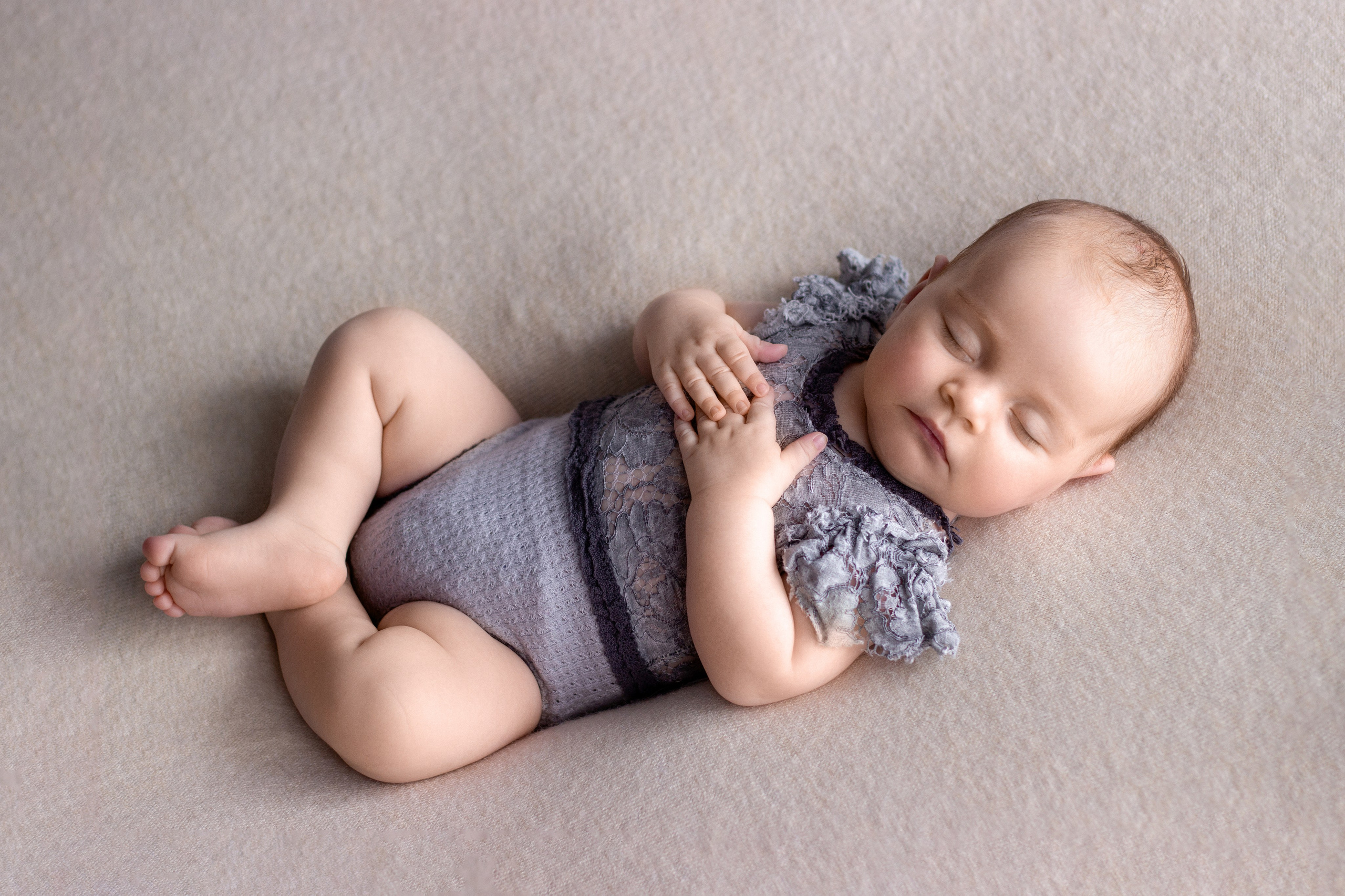 спящая капризная новорожденная белокурая малышка лежит на спинке в нежном сером боди с рюшами на теплом бежевом фоне