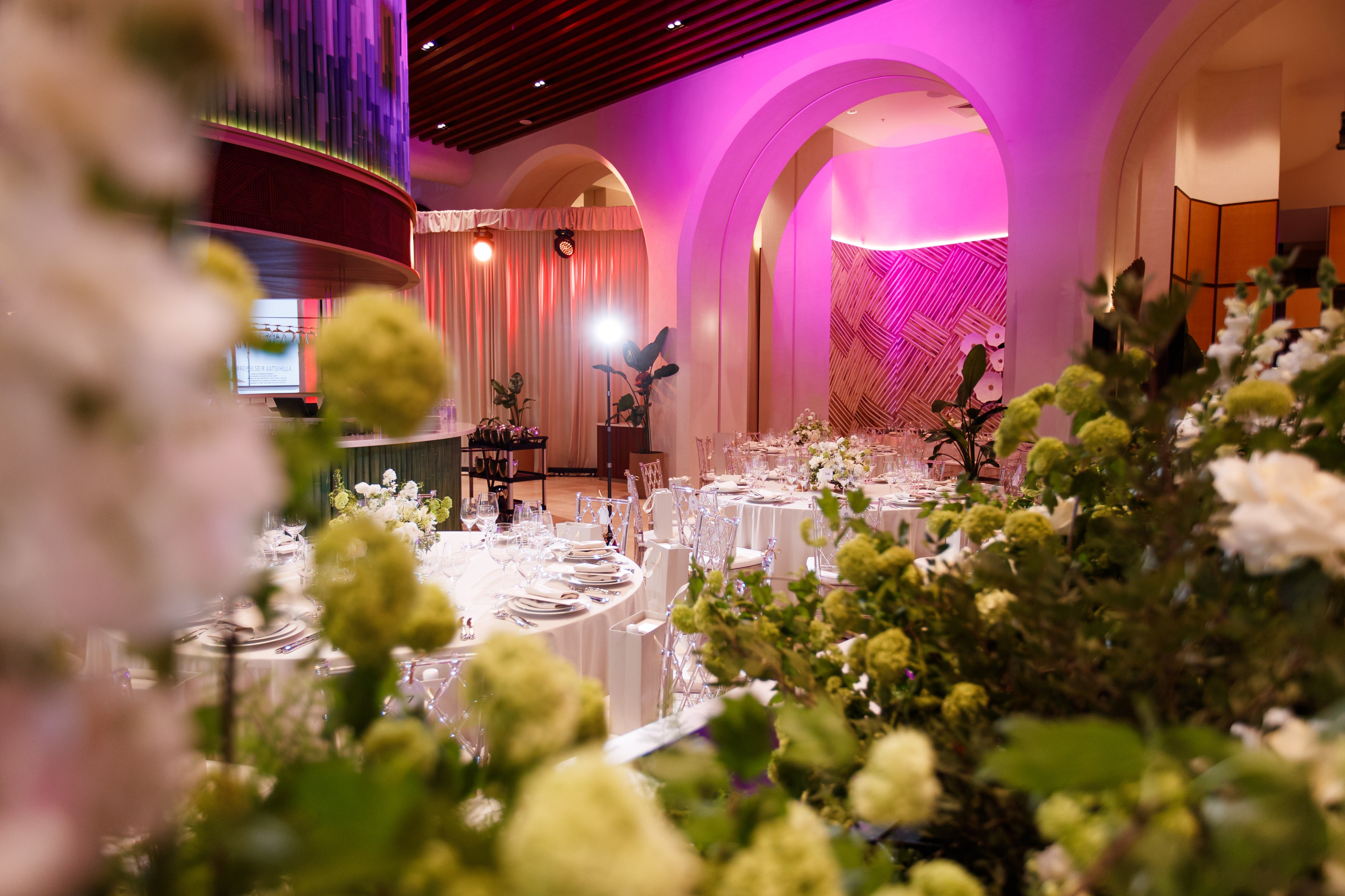 Банкетный зал: Вид на праздничные столы через пышные букеты цветов в интерьере с розовым освещением.