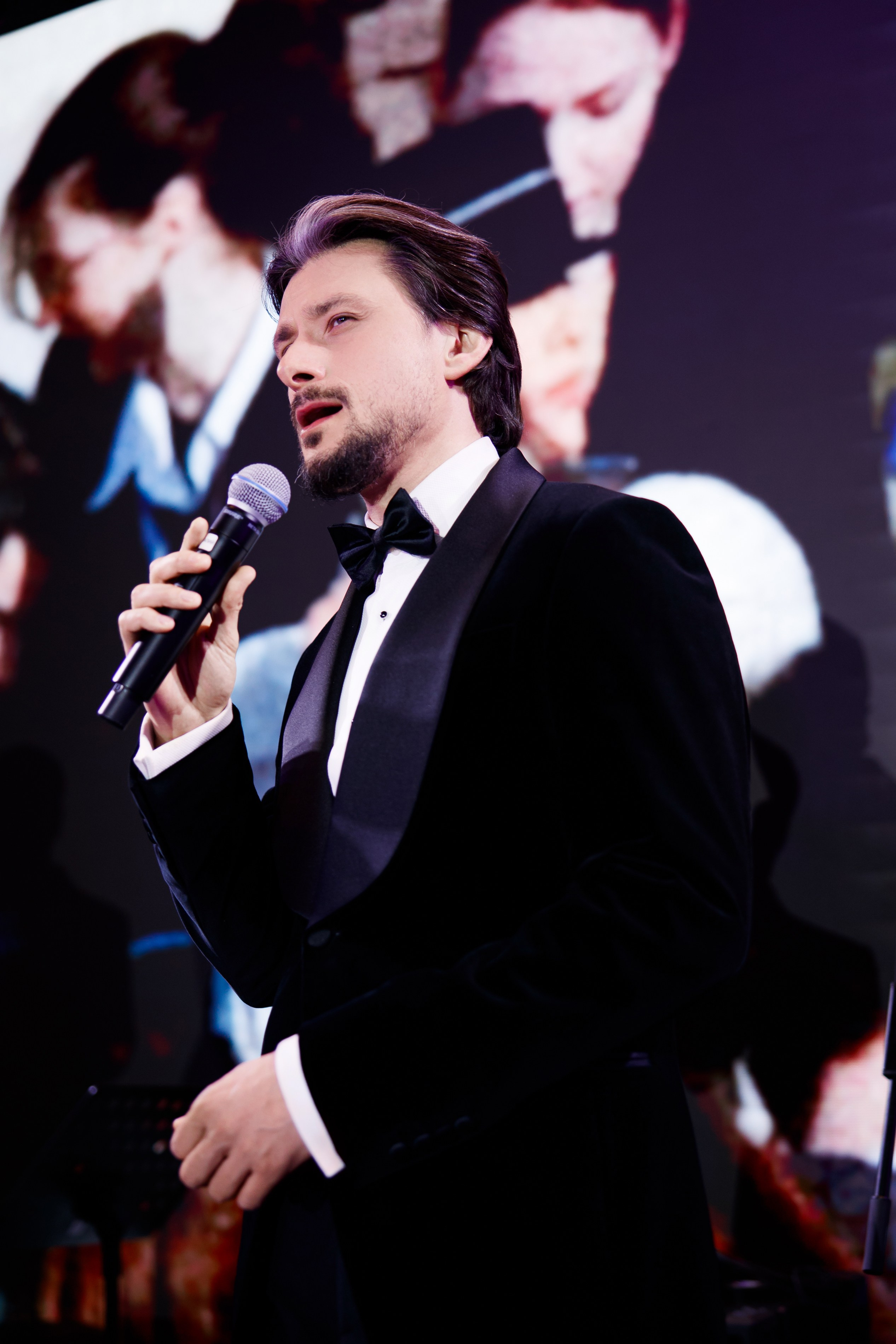 Репортажная съемка ведущих на светском мероприятии: ведущий в классическом черном смокинге с бабочкой на фоне вечернего зала.