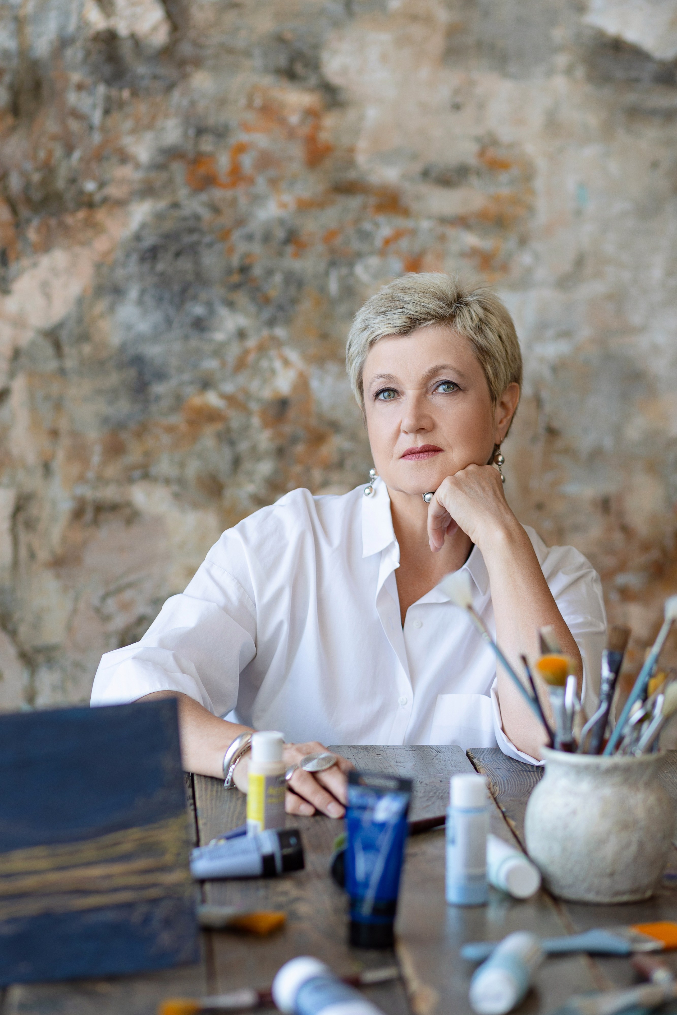 Женщина сидит за столом . На столе ваза с кистями, тюбики с красками, написанный холст.