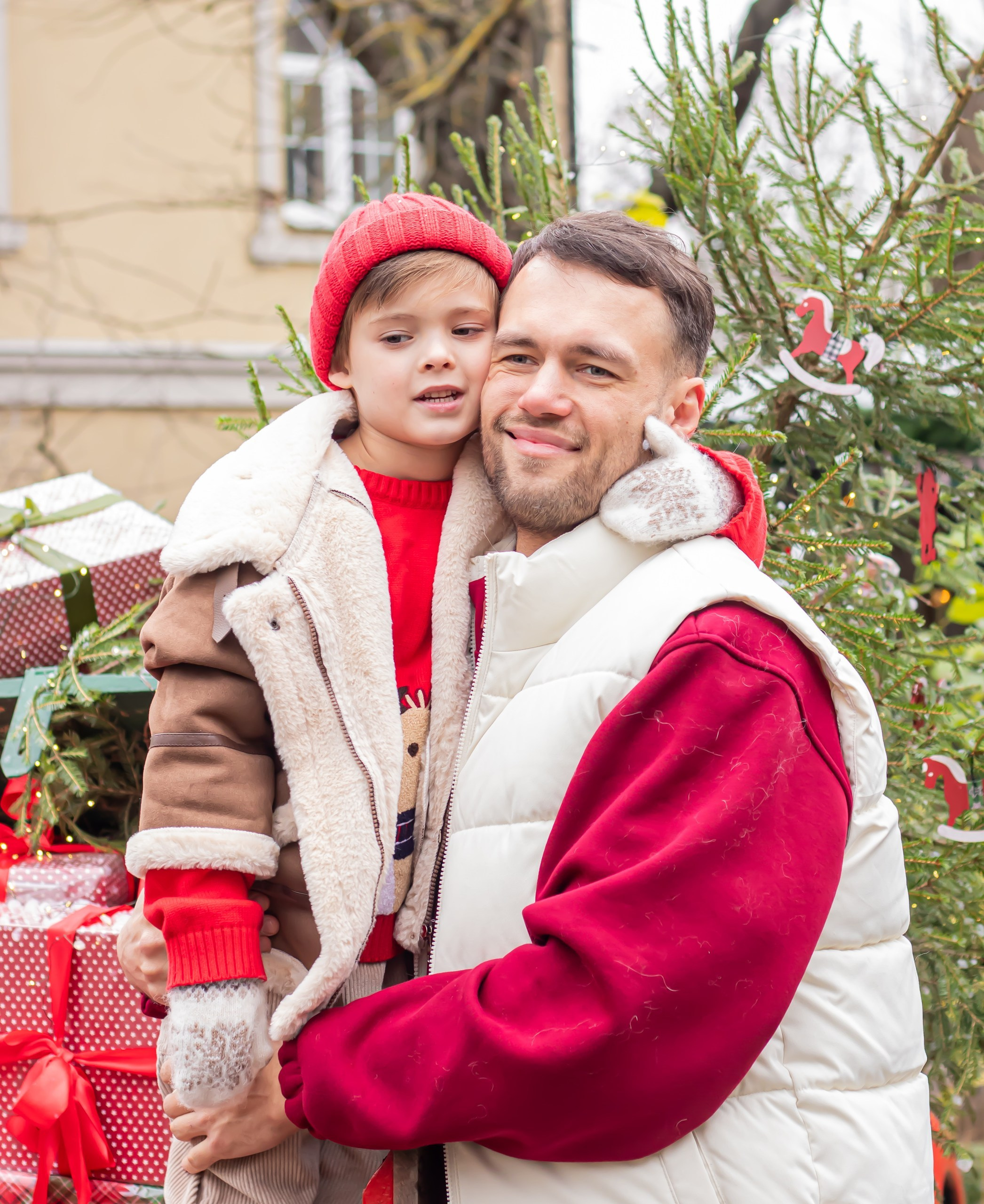 Папа и сын в красно-белых костюмах улыбаются, стоя у новогоднего декора с ёлками, фотограф Карасёва Елена