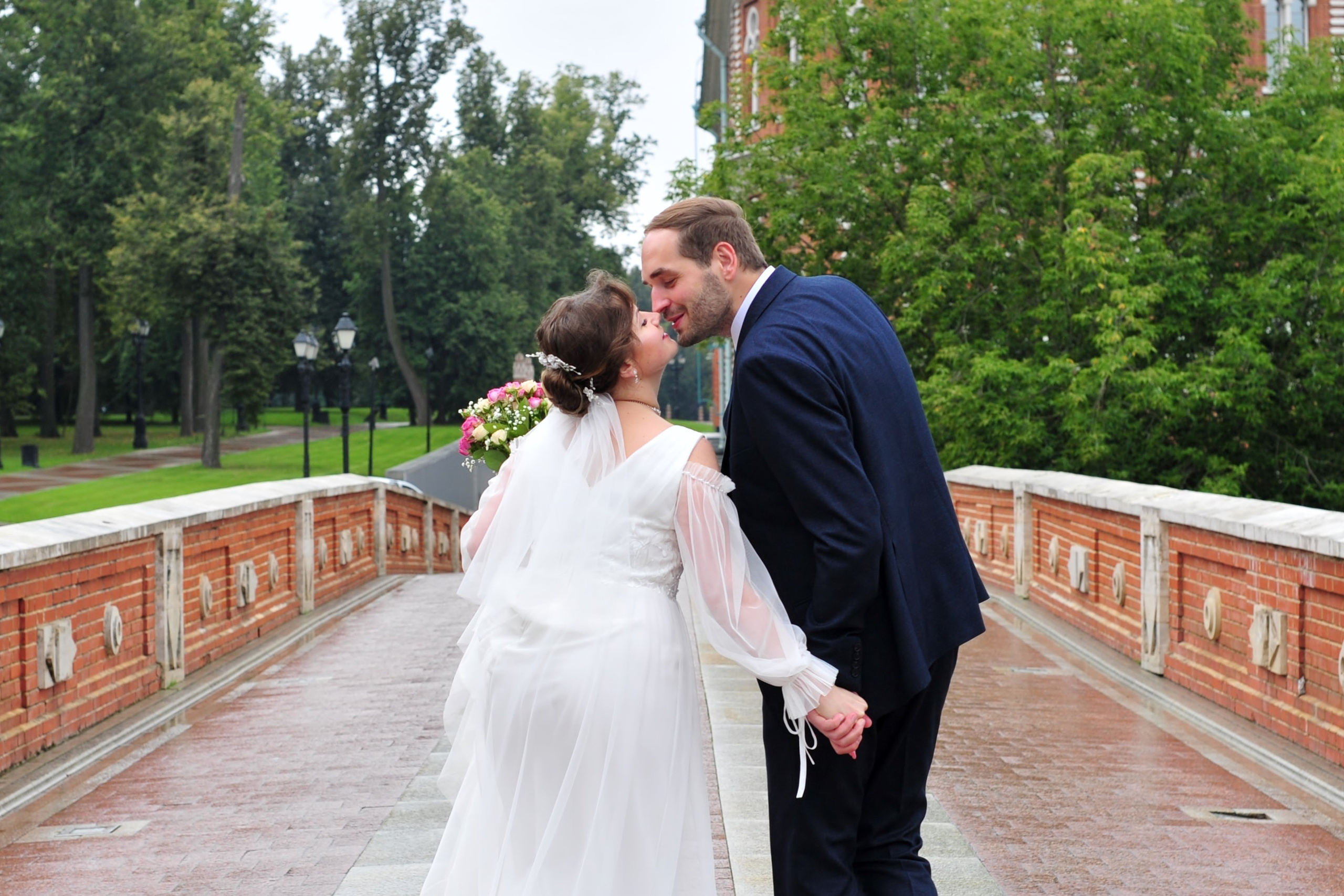 Свадьба в Царицыно пасмурным летним днем, поцелуй новобрачных, вид со спины на каменном мосту из красного кирпича 