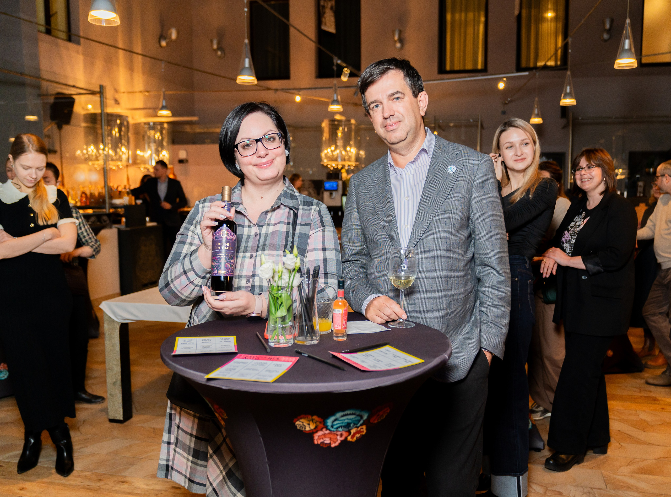 Женщина в клетчатой рубашке держит бутылку вина «Медовые слезы» и улыбается вместе с мужчиной в сером пиджаке на дегустации в Radisson Sonya. Фотография Евгения Мальцева (evrednix.ru).