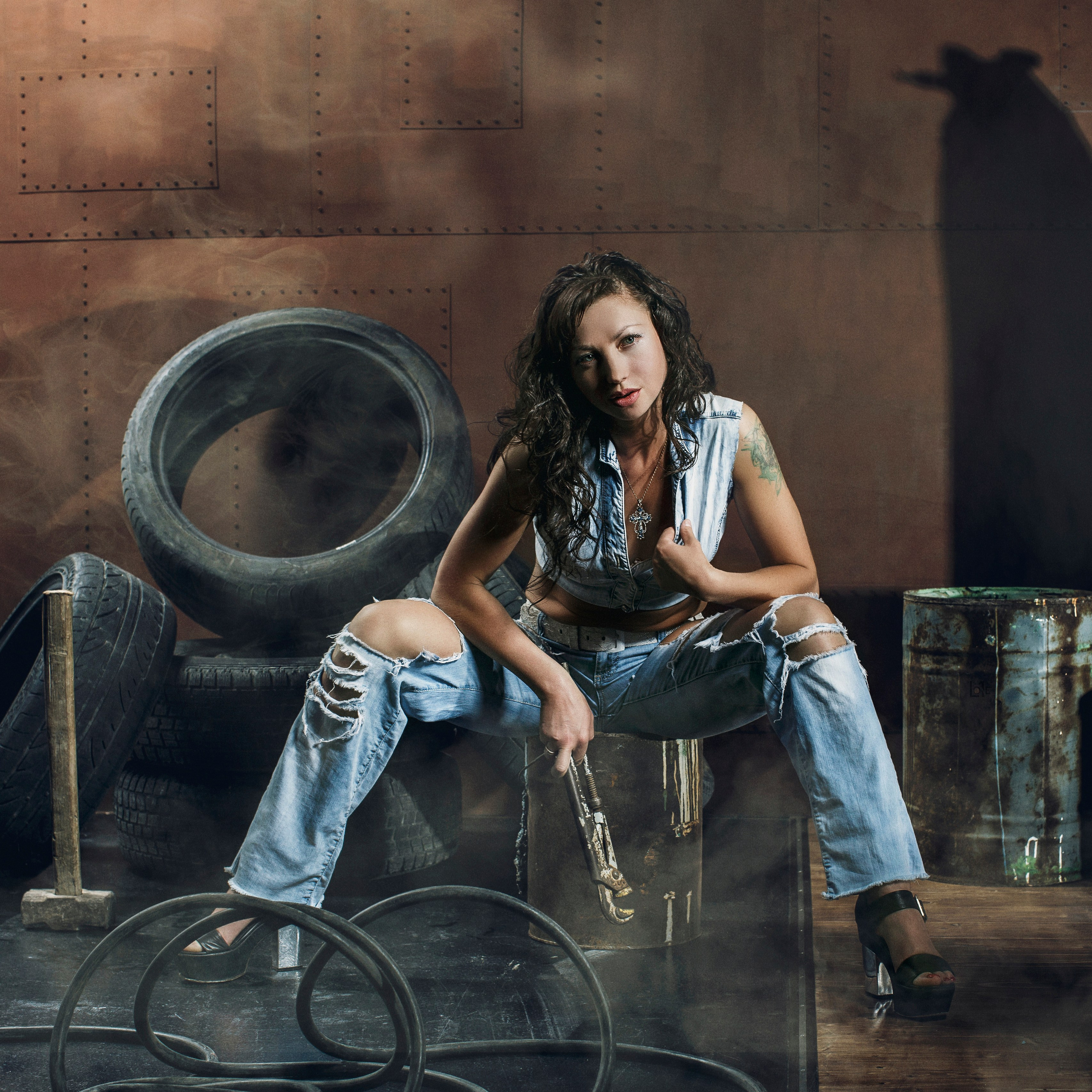 девушка слесарь в гараже сидит на бочке в рваных штанах с ключом в руке на заднем фоне автомобильные покрышки