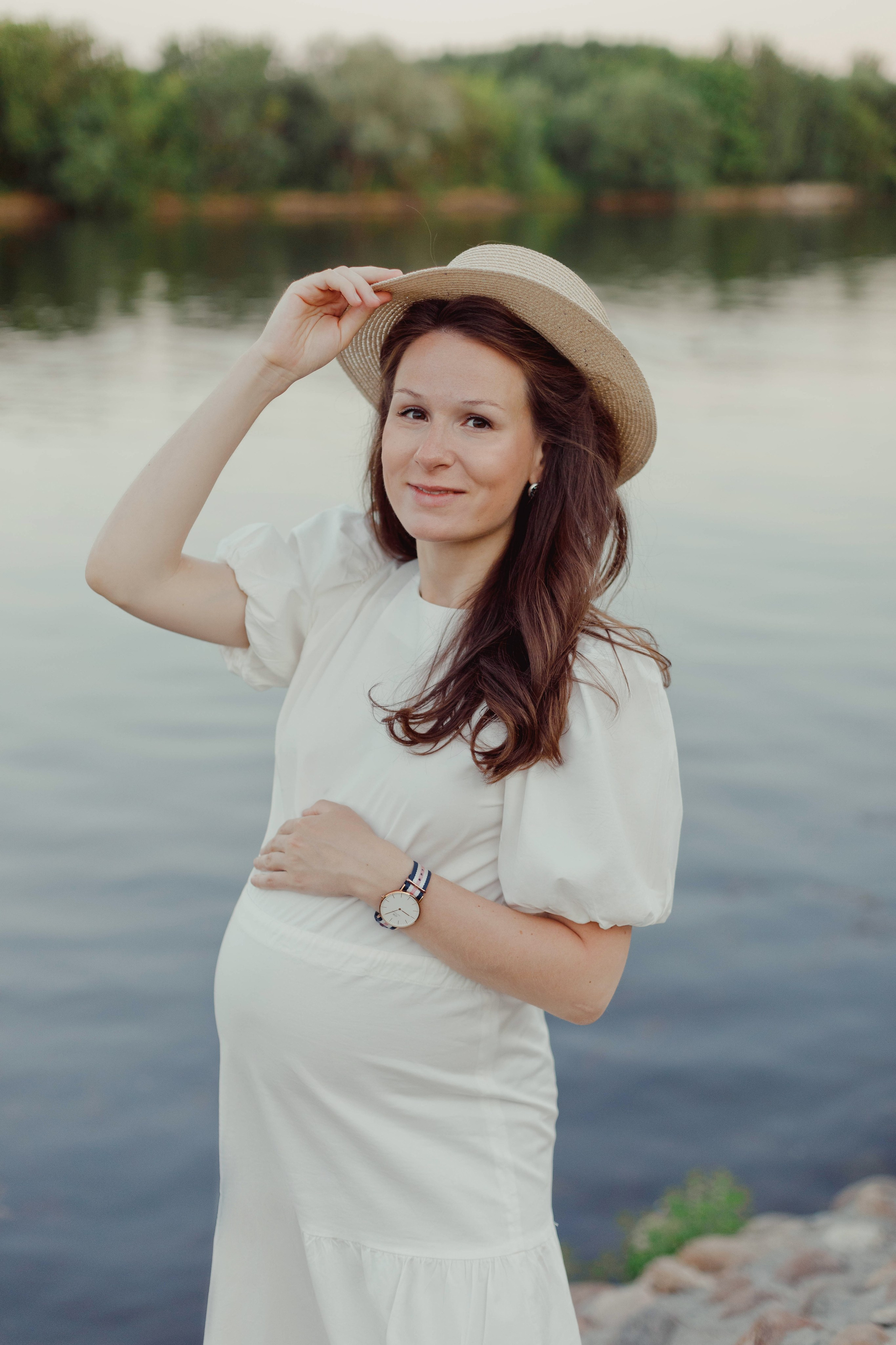 Романтический портрет беременной женщины в летнем платье и шляпе на берегу реки. Съёмка беременности на природе.