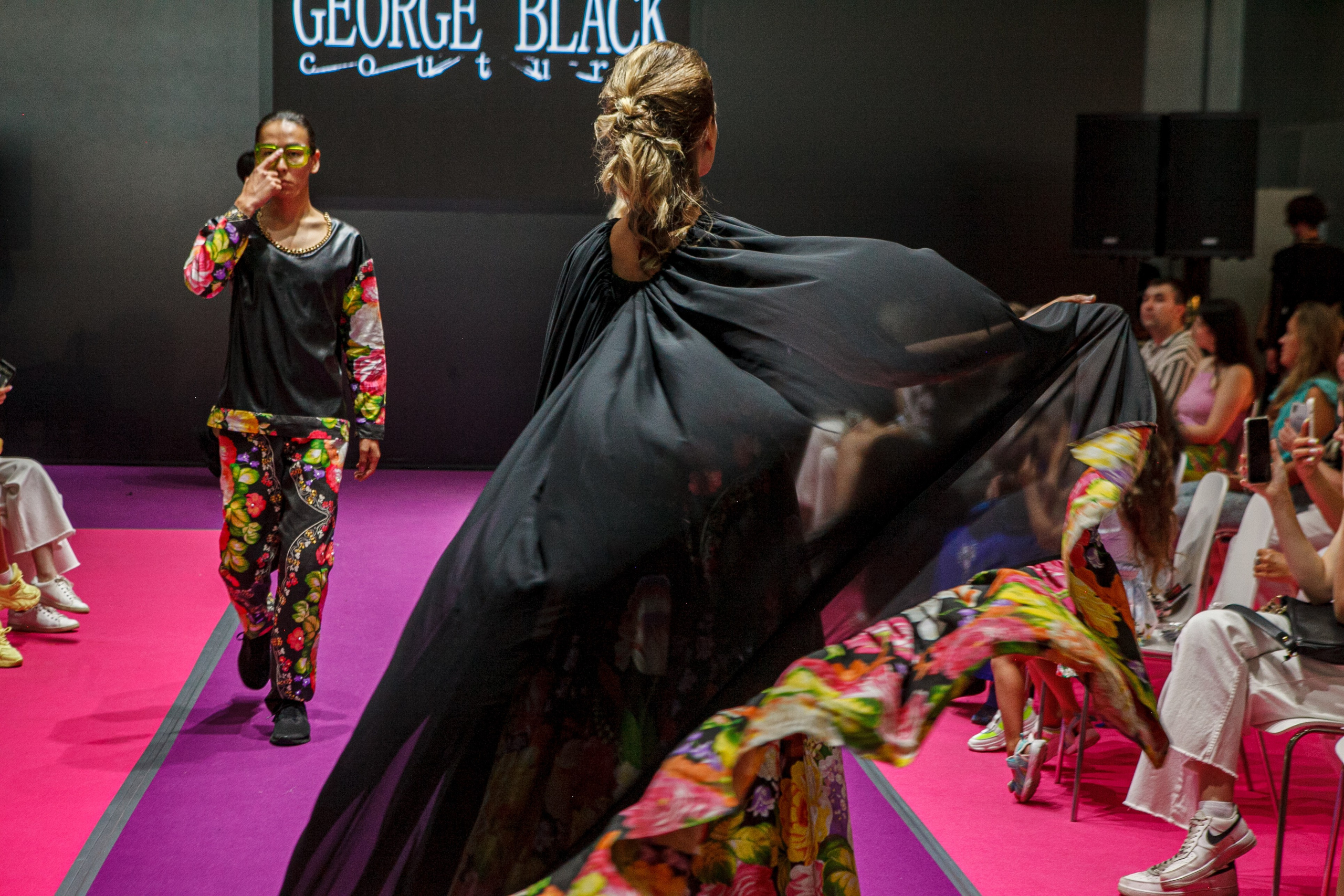  "Модели в ярких нарядах проходят по подиуму во время показа мод, освещенные яркими софитами и окруженные восторженными зрителями."