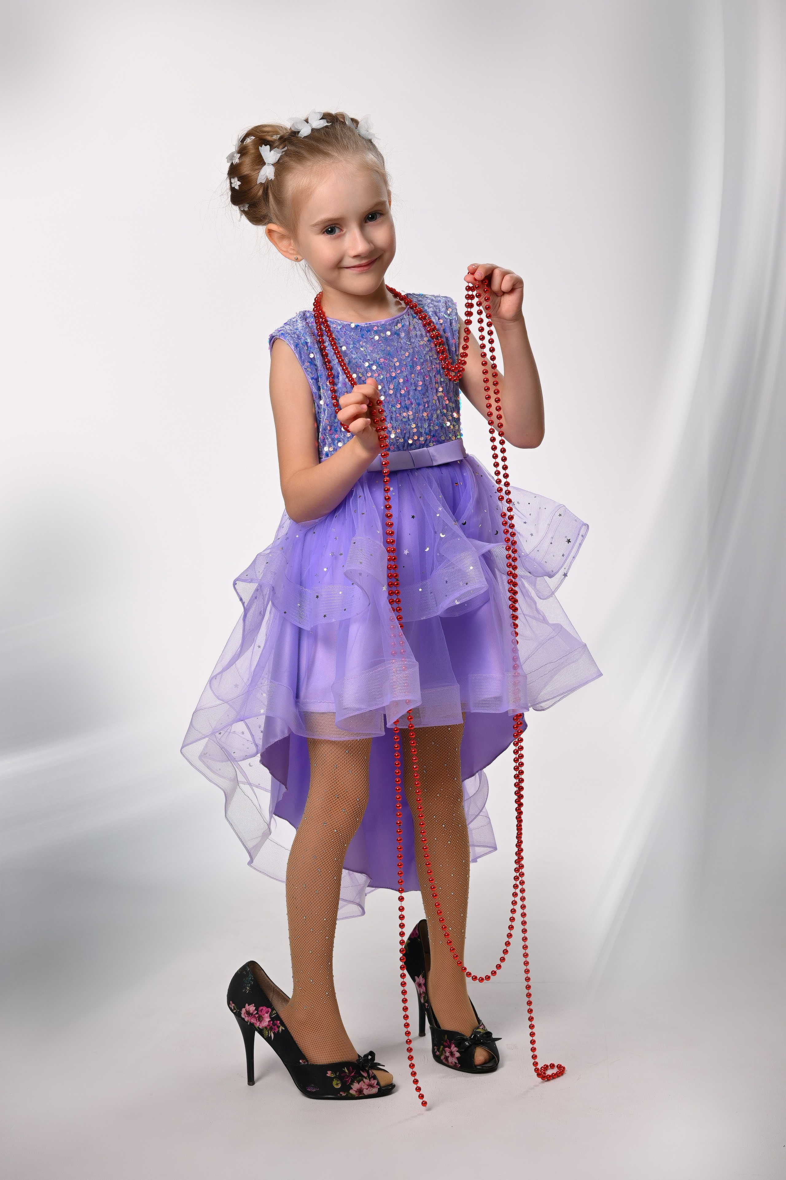 Девочка в выпускном платье и больших туфлях держит длинные бусы в фотостудии