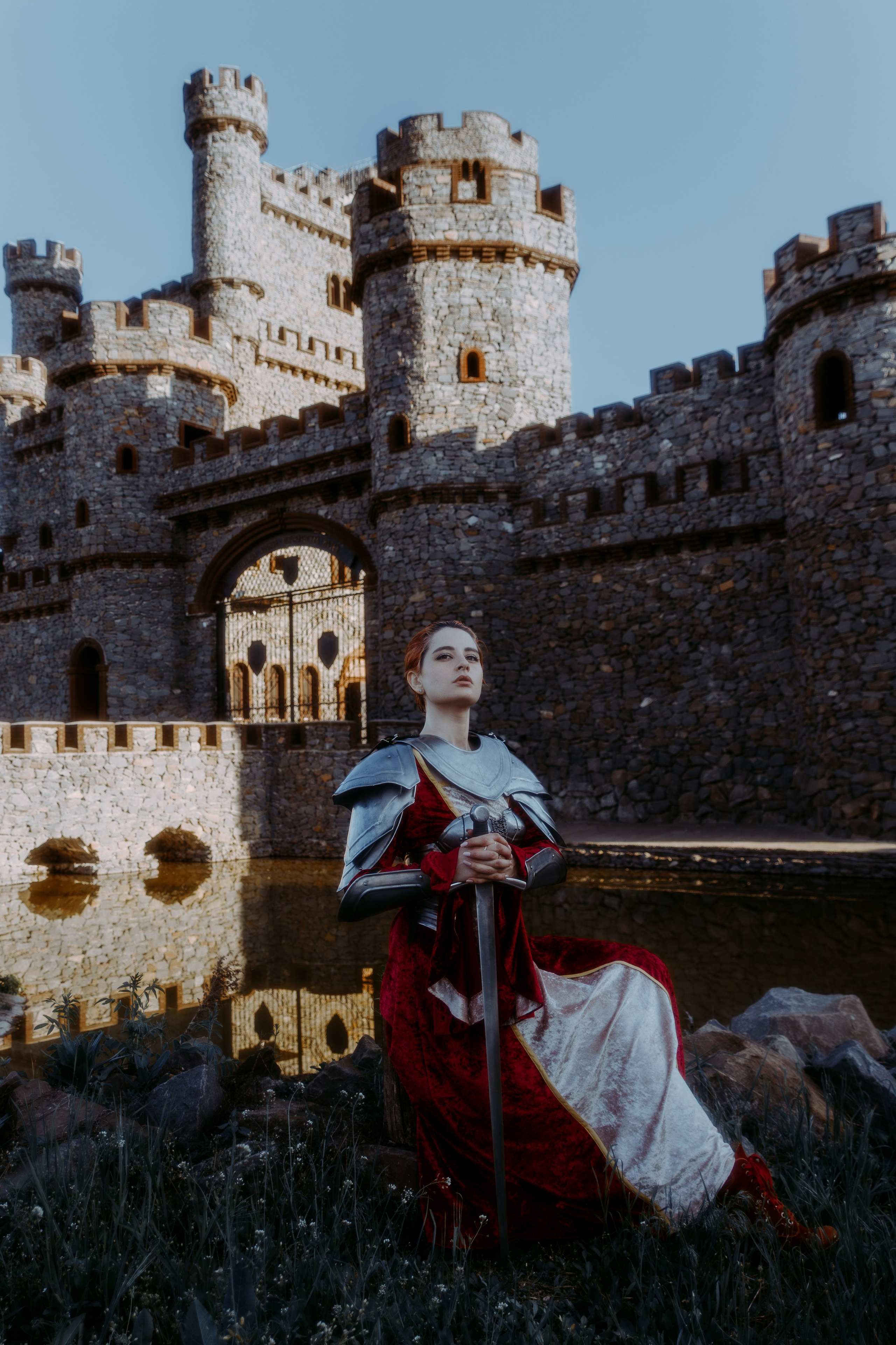 Девушка в рыцарских доспехах сидит на камне на фоне замка и воды