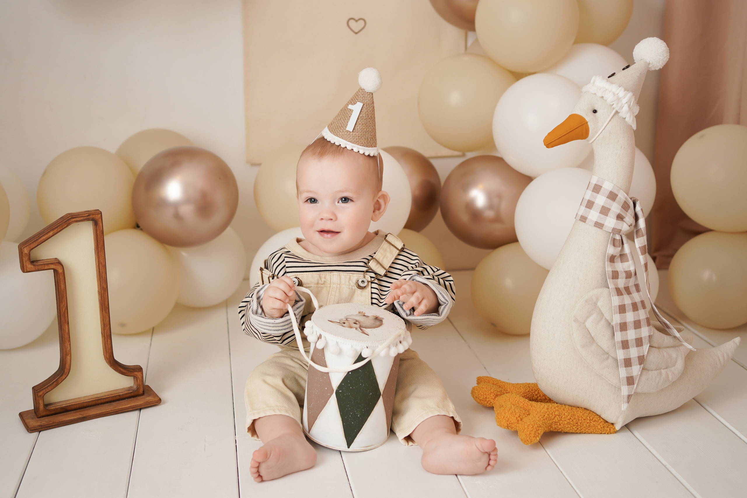 На фото малыш в колпачке, рядом единица, игрушка гусенок, много шаров в бежевой гамме.
