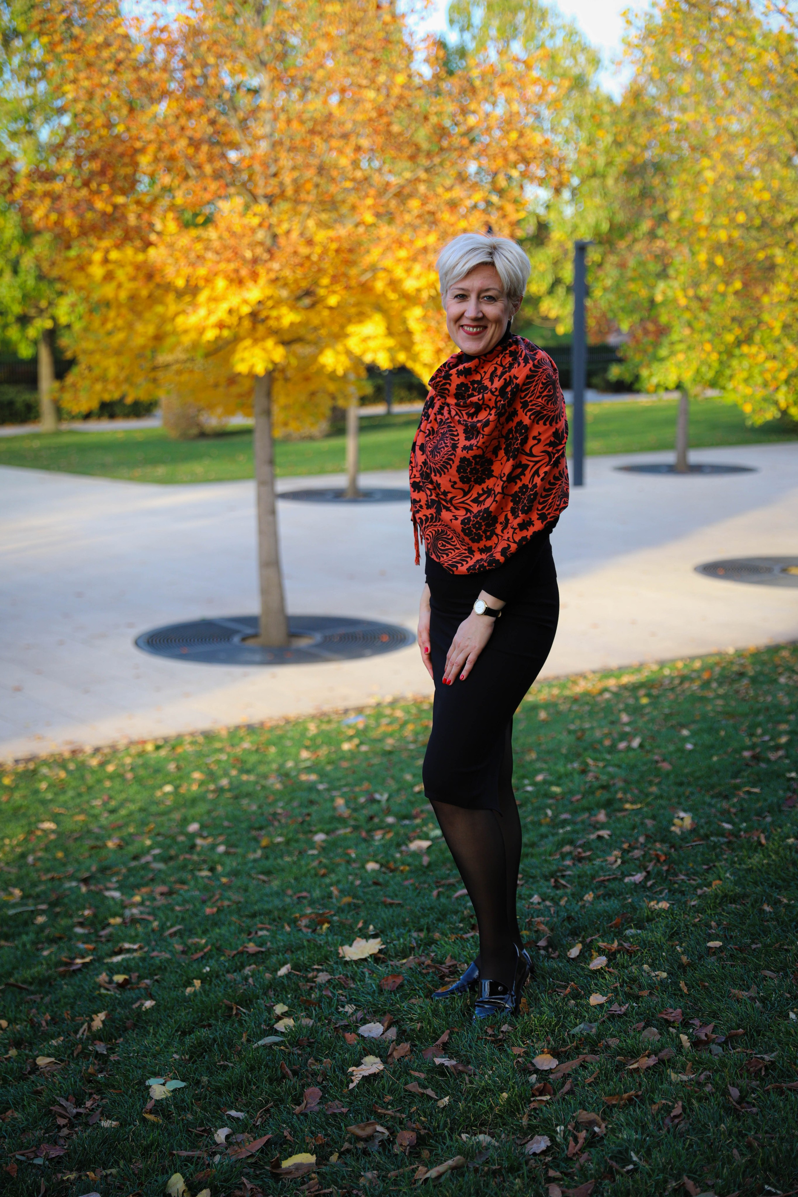 Картинка осени: молодая женщина в оранжевом платке проходит мимо деревьев, усыпанных желтыми листьями.