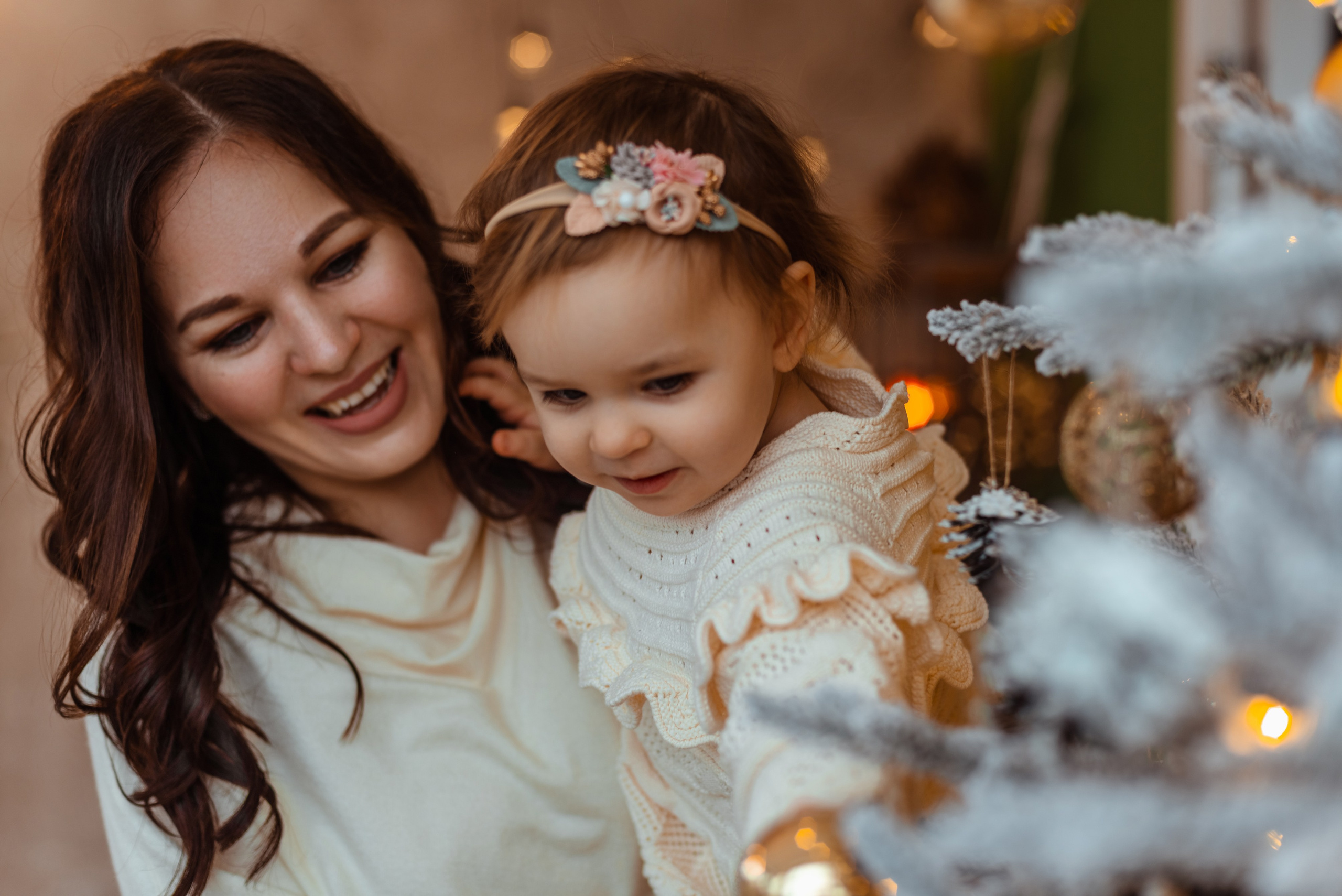 Мама в белом платье улыбается, держа на руках маленькую дочку в светлом платье с кружевами. Они находятся рядом с украшенной новогодней елкой.