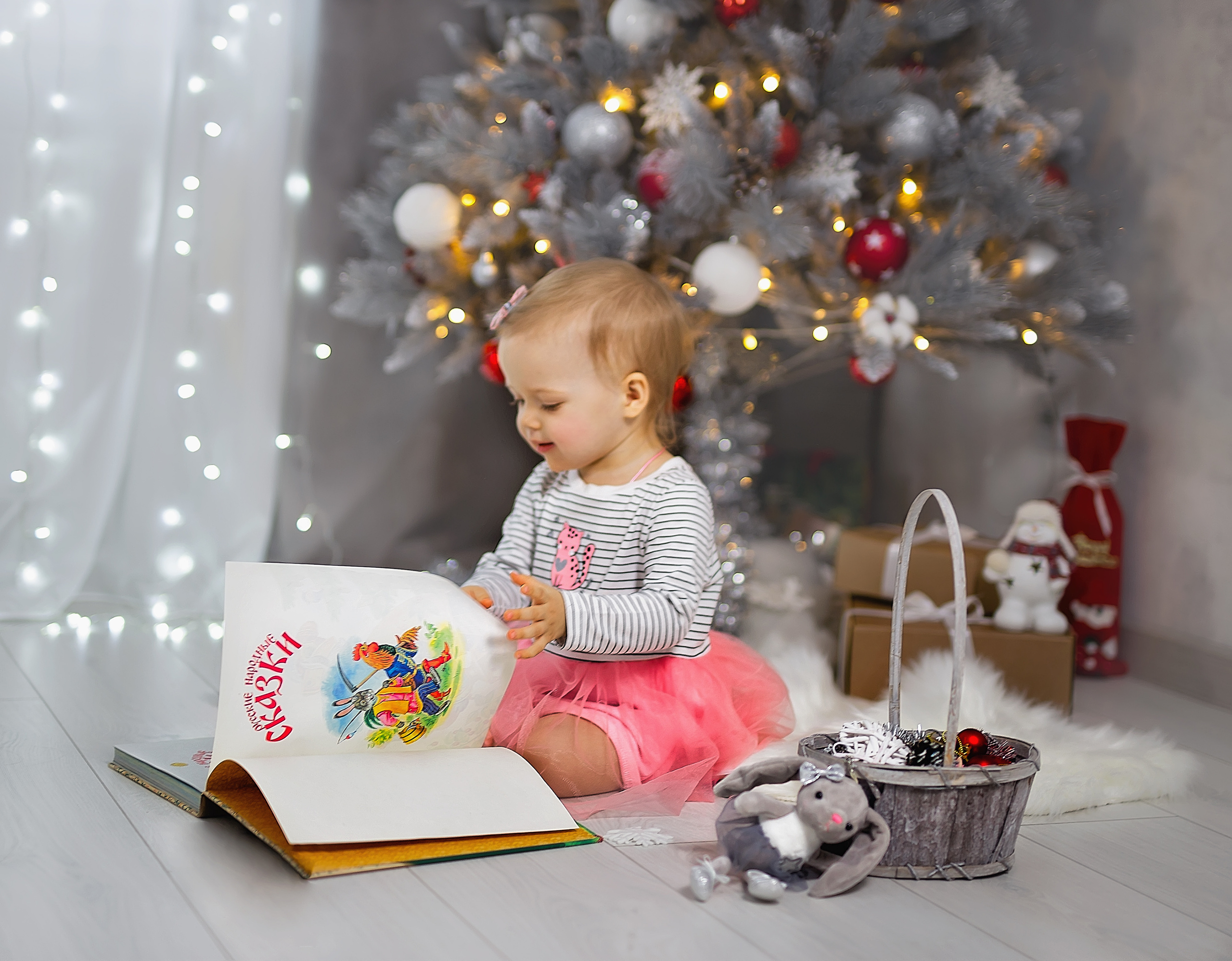 Девочка листает книжку со сказками. Сзади горят огоньки и стоит красивая серая елка.