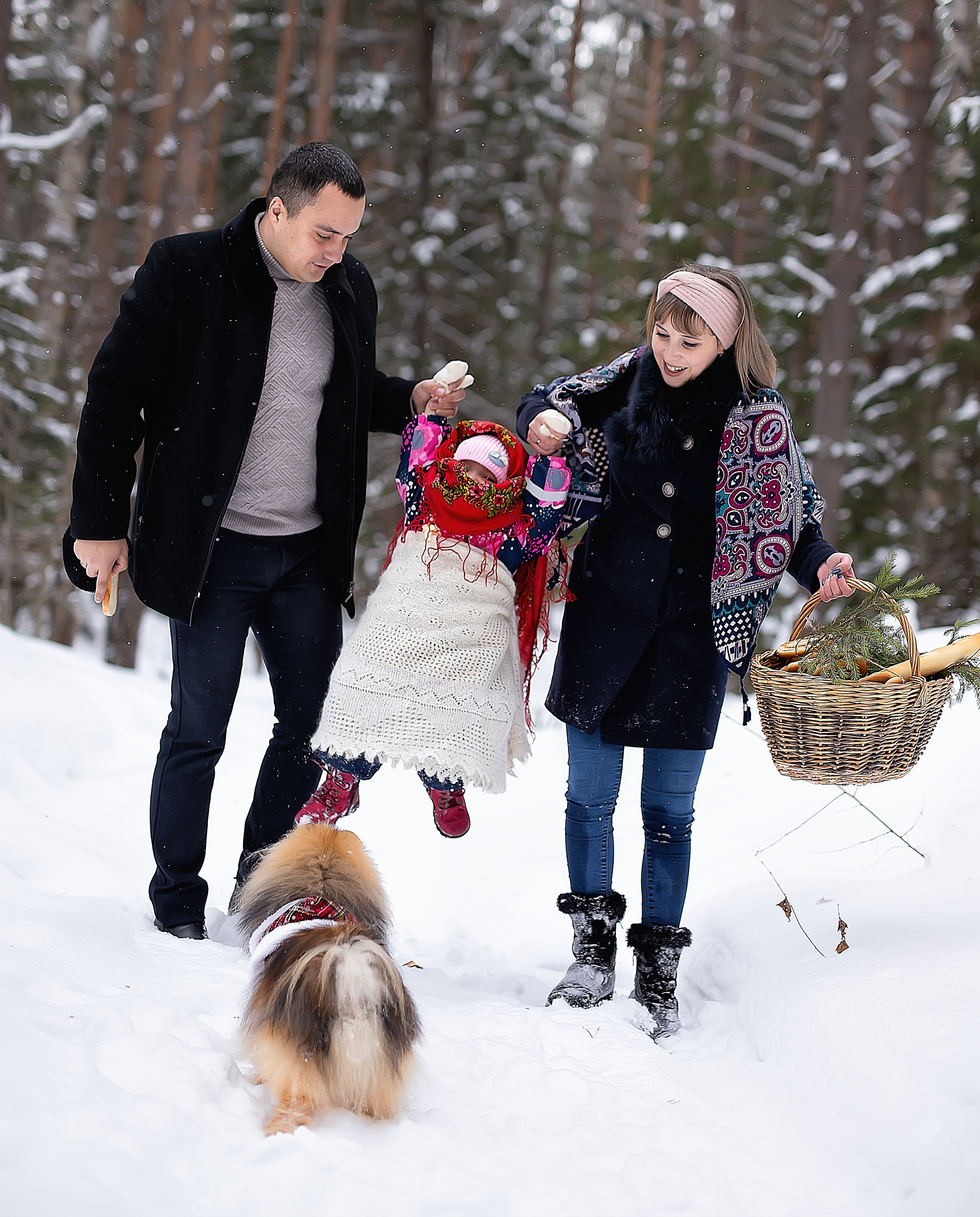 Счастливая семья гуляет по лесу поднимая ребенка в воздух, пока собака за всем этим наблюдает.