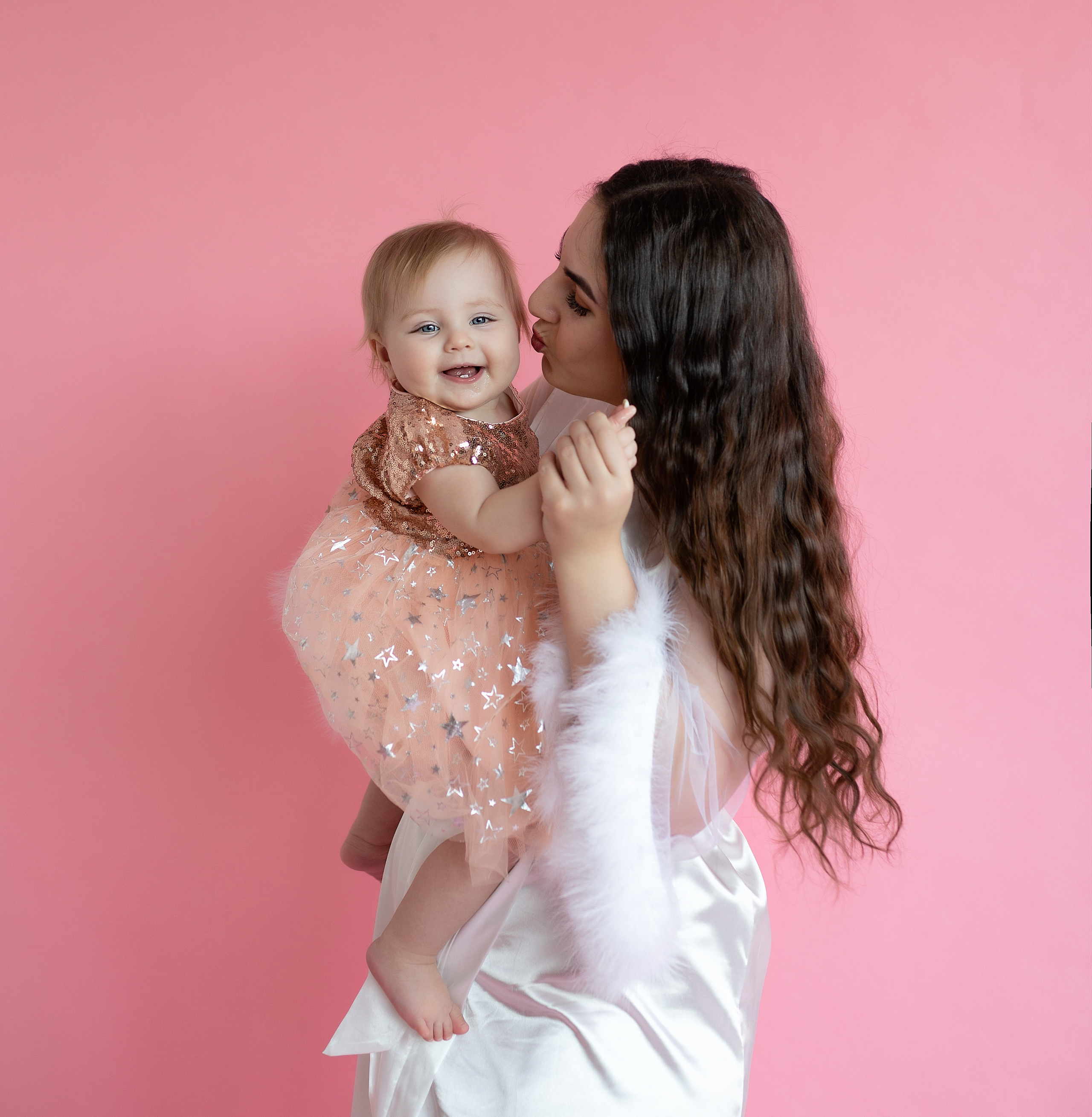 Женщина целует своего ребёнка в щечку. Она одета в белое платье.