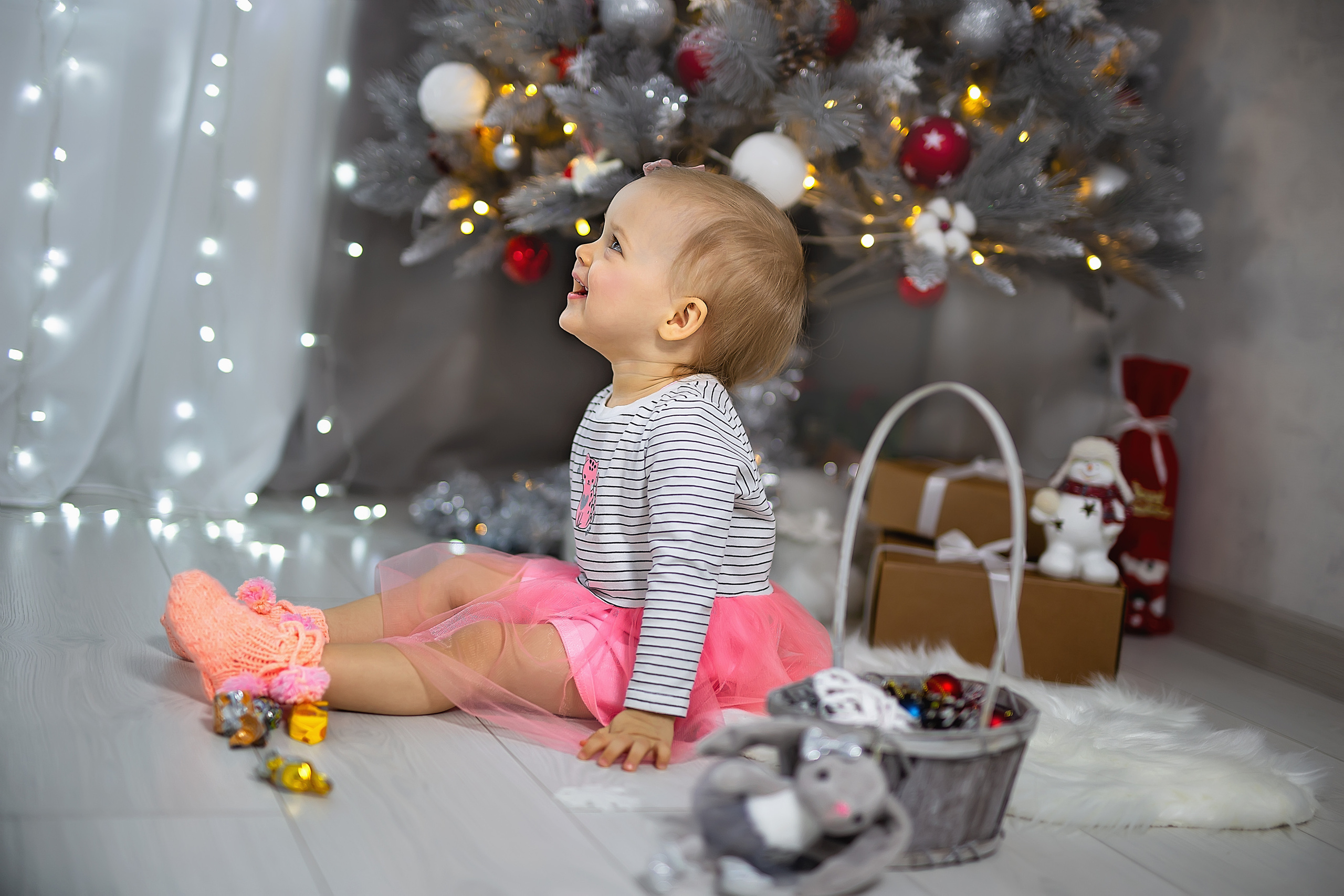 Девочка сидит под елочкой перед Новым годом в красивой розовой юбке и смеется 