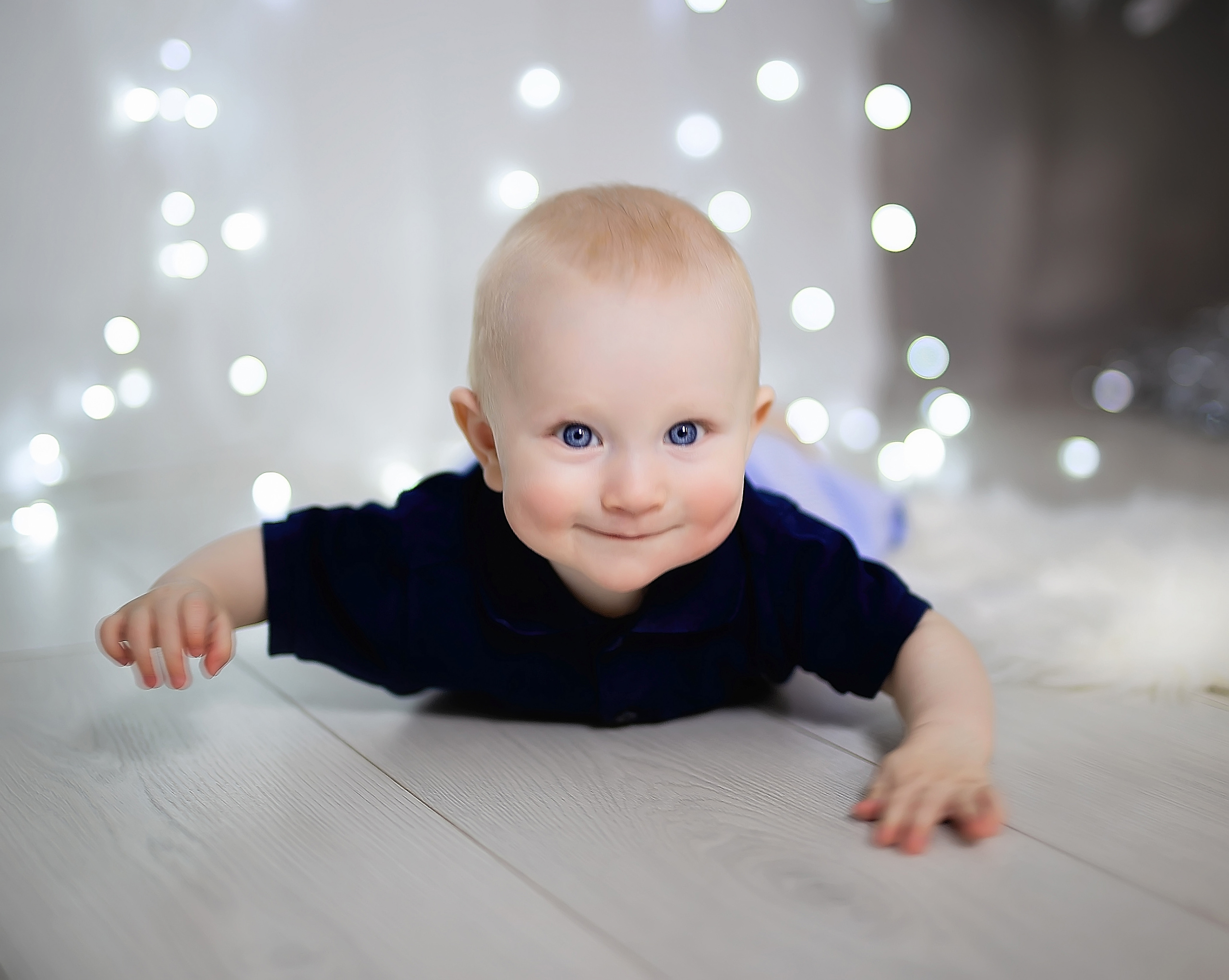 Счастливый малыш с красивыми глазами лежит на животе и улыбается. Сзади горят фонарики.