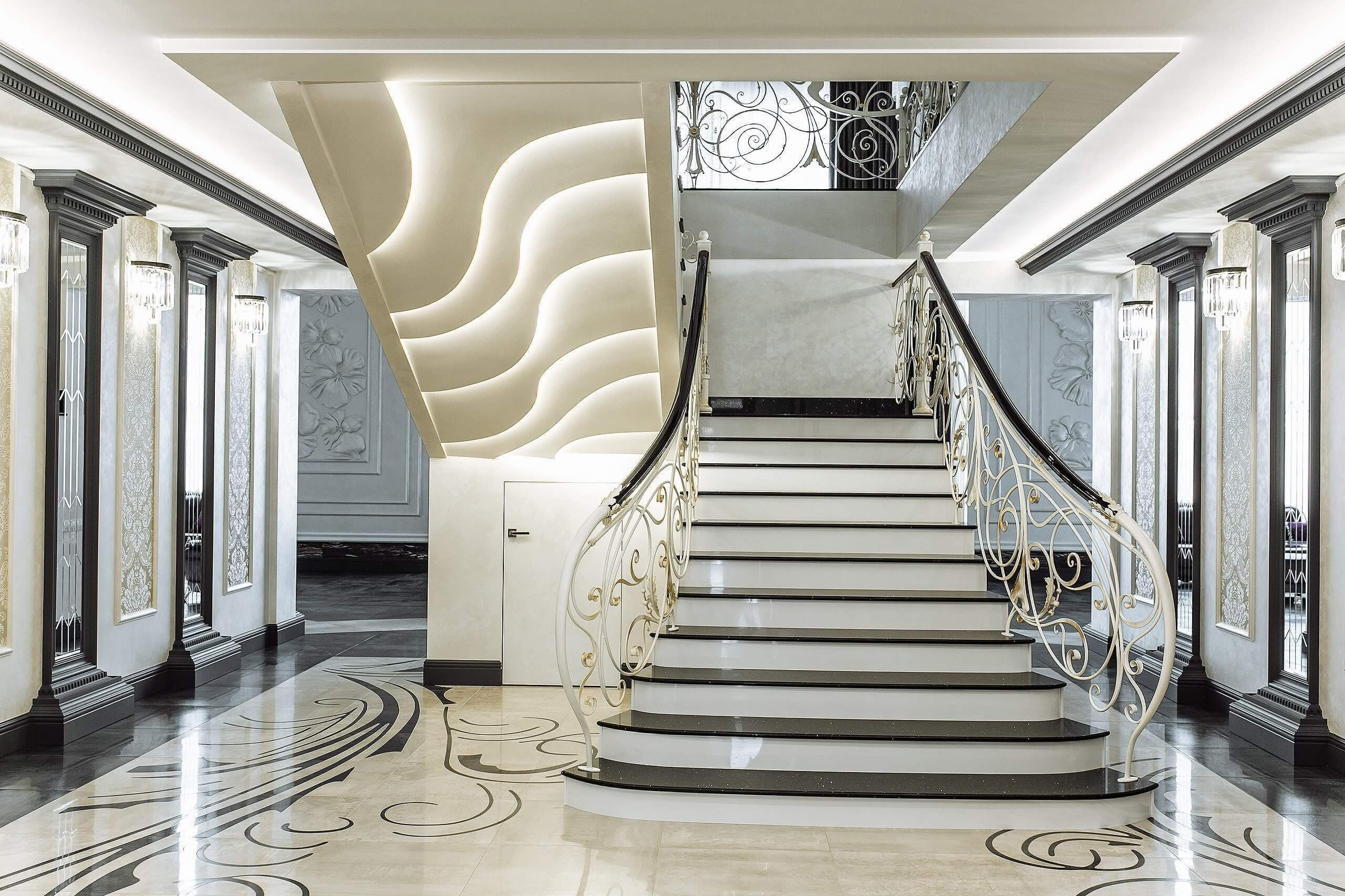 Фотография дизайна холла в стиле современная классика: лестница с черно-белыми ступенями и ажурными коваными перилами, скульптурная 3D-панель с подсветкой и мраморный пол с графическим рисунком. Снято для проекта загородного дома в Пензе.