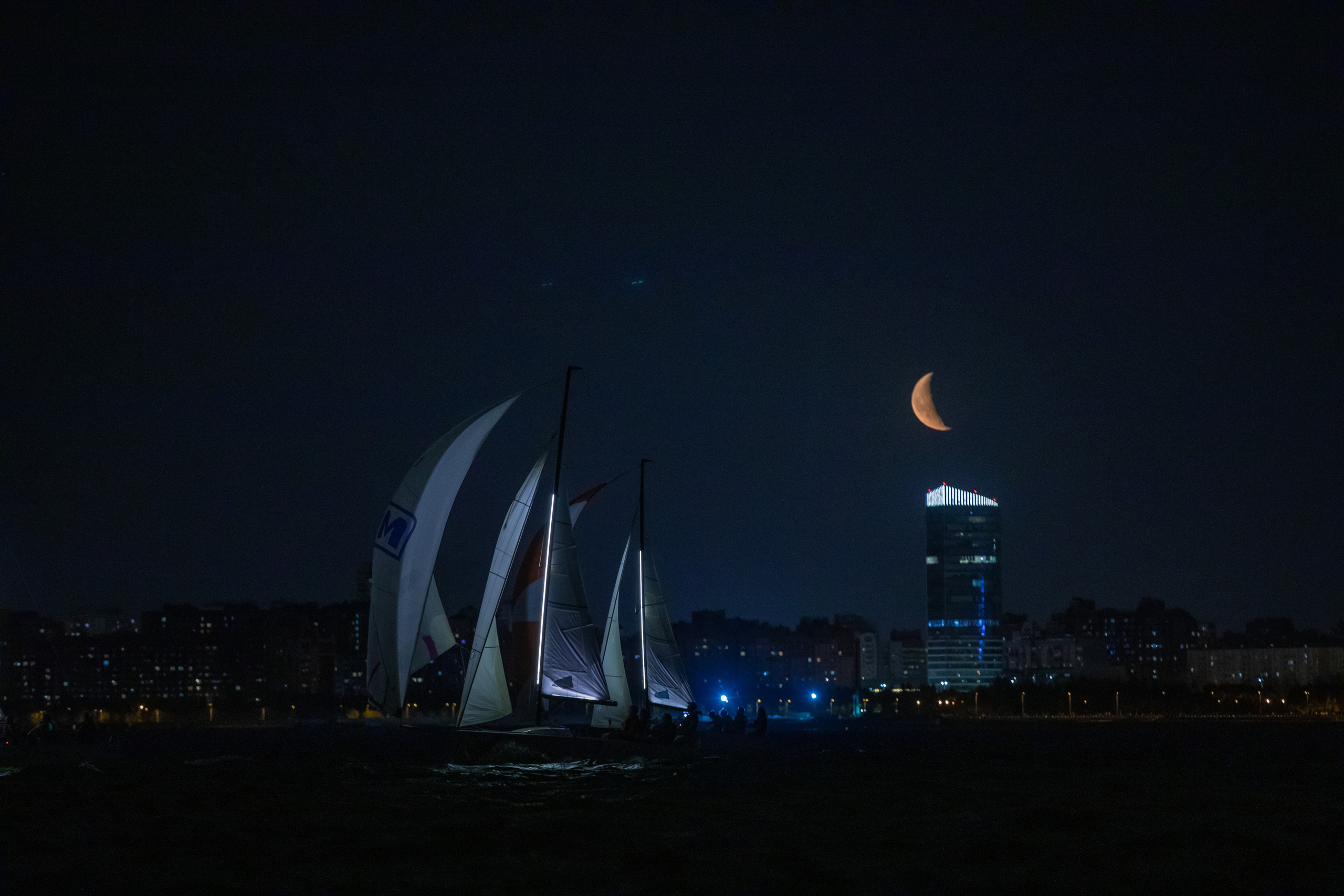 Парусные яхты идут по Финскому заливу на фоне Атлантик Сити и луны. Ночная регата в Санкт-Петербурге.