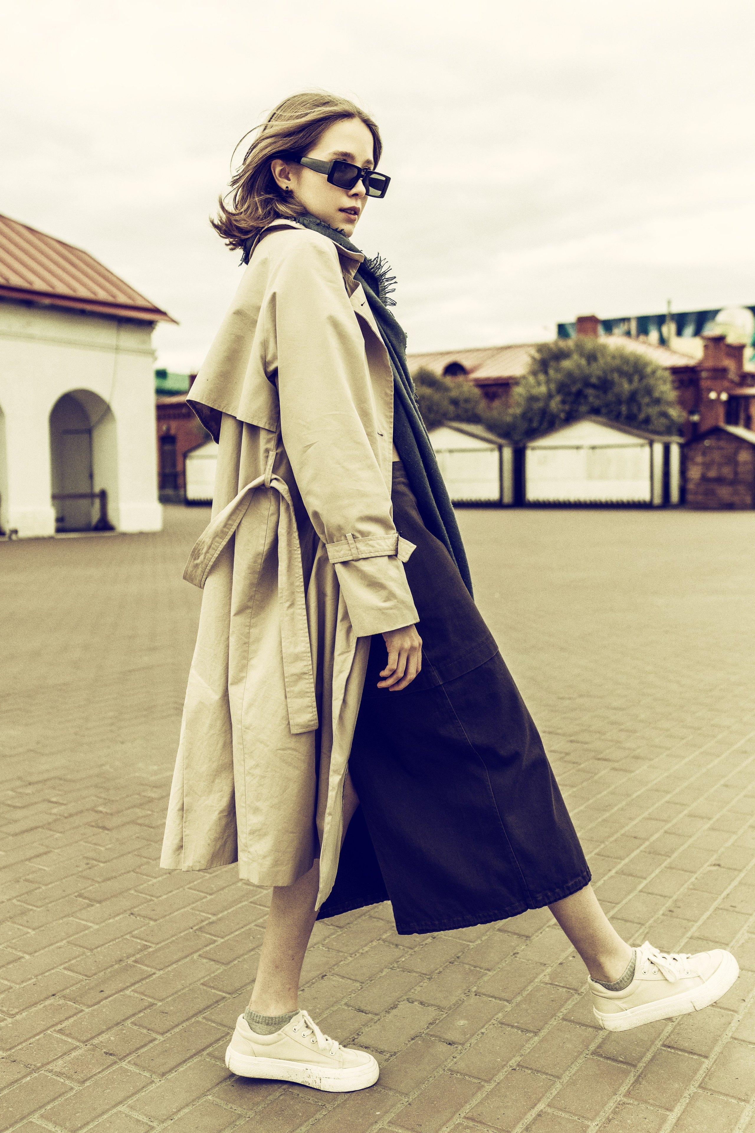 Девушка  в бежевом плаще и солнцезащитных очках уверенно шагает по мощеной  площади. Пасмурное небо и приглушенные тона создают стильное, непринужденное настроение.