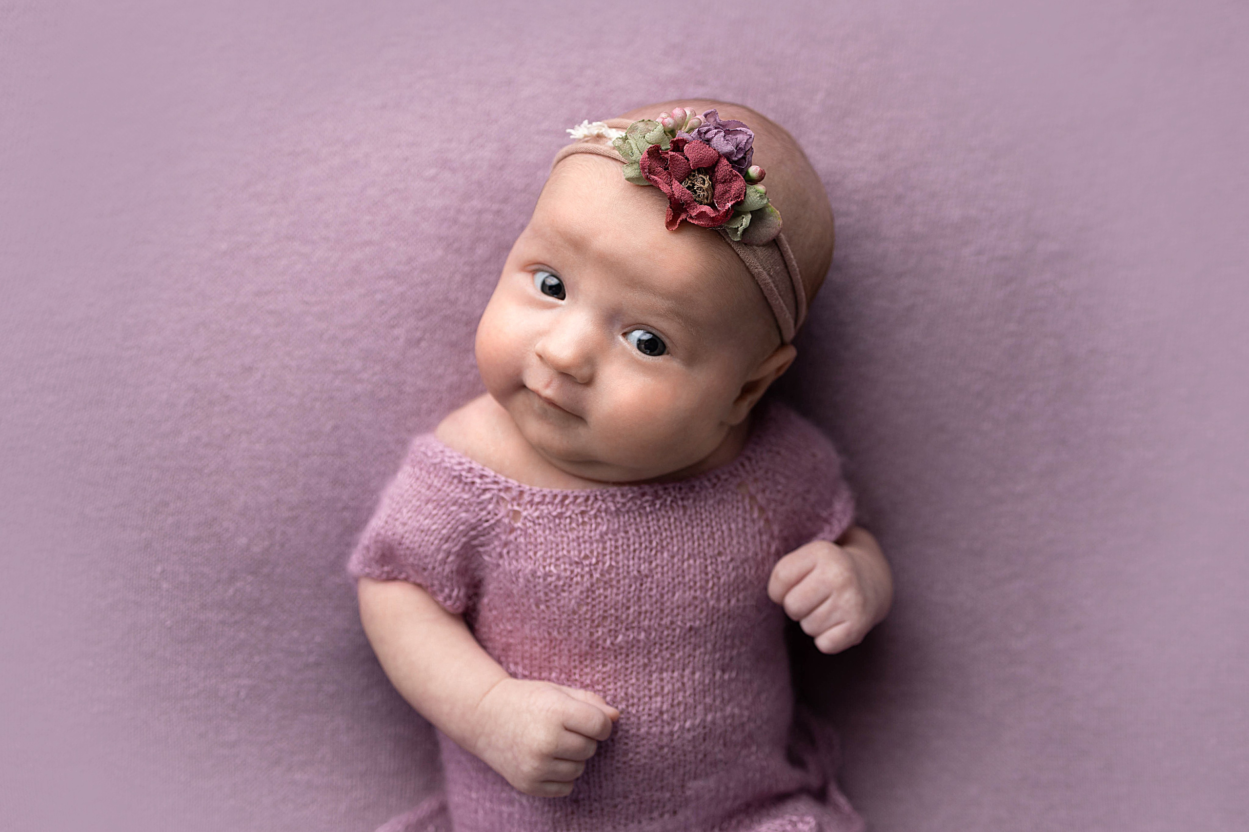 Улыбающийся новорожденный в пурпурном наряде с цветочным украшением на голове.