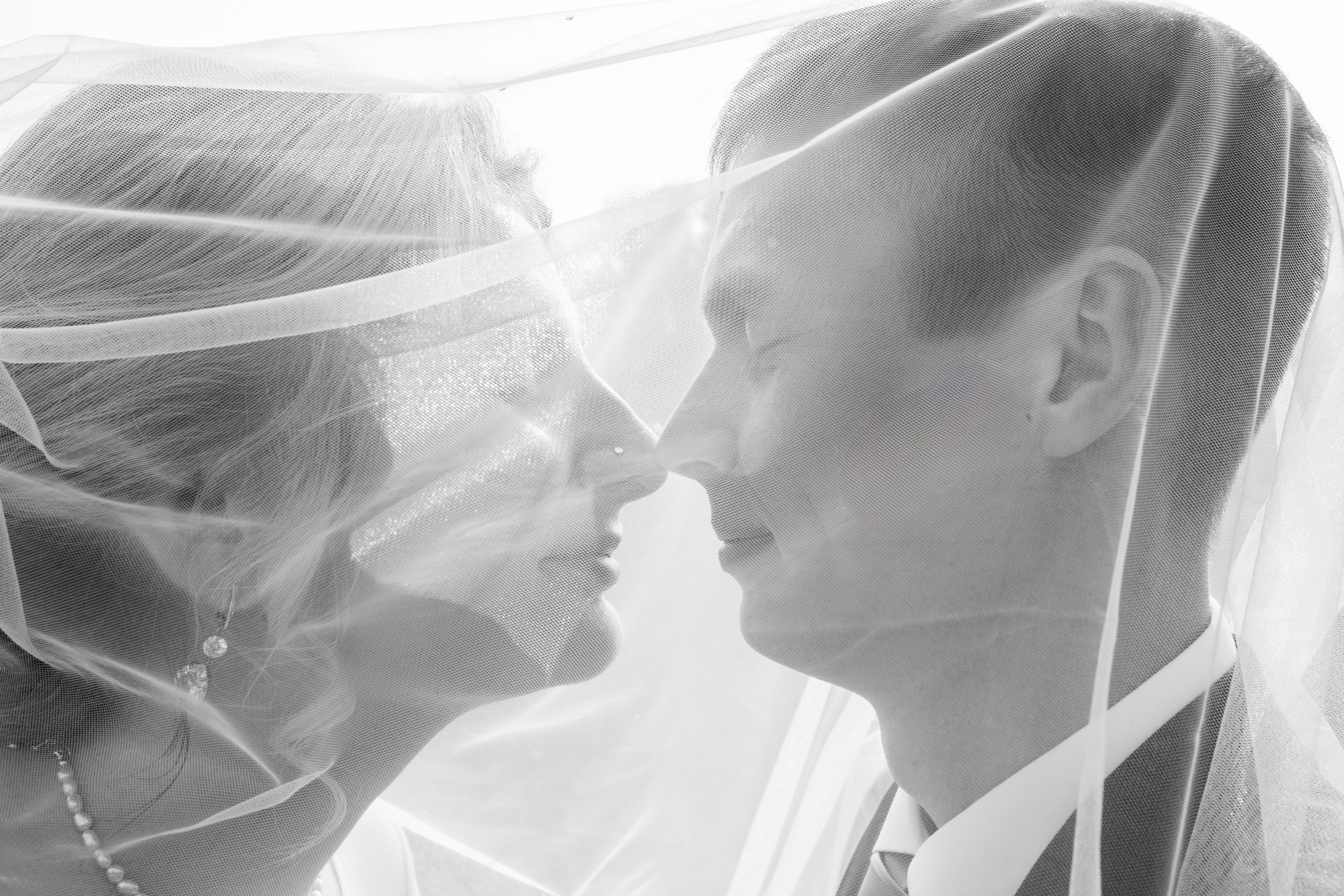 Романтичный момент под фатой — жених и невеста смотрят друг на друга в полупрозрачном свете.