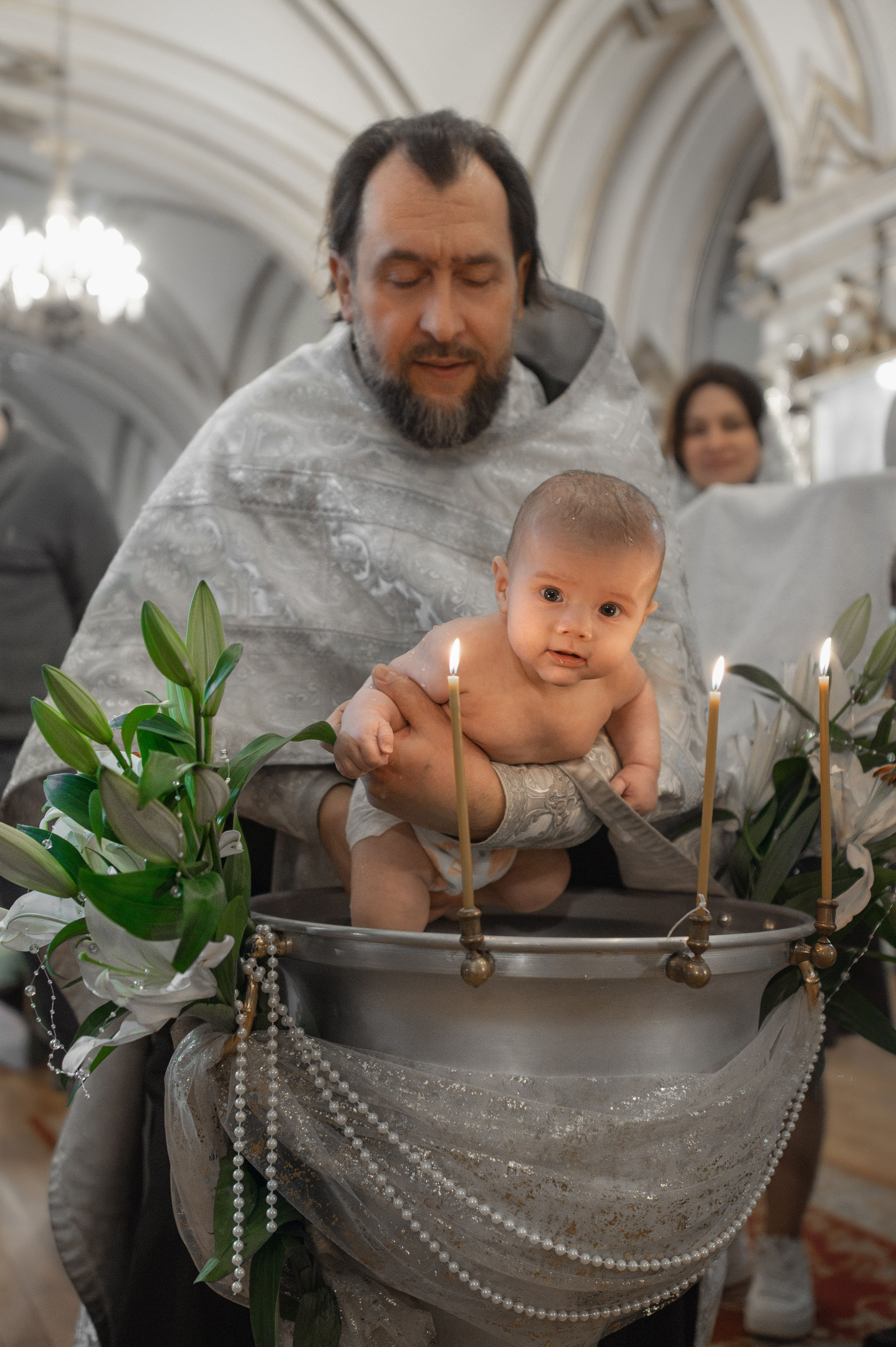 священник сажает малыша в украшенную цветами купель Никольского собора