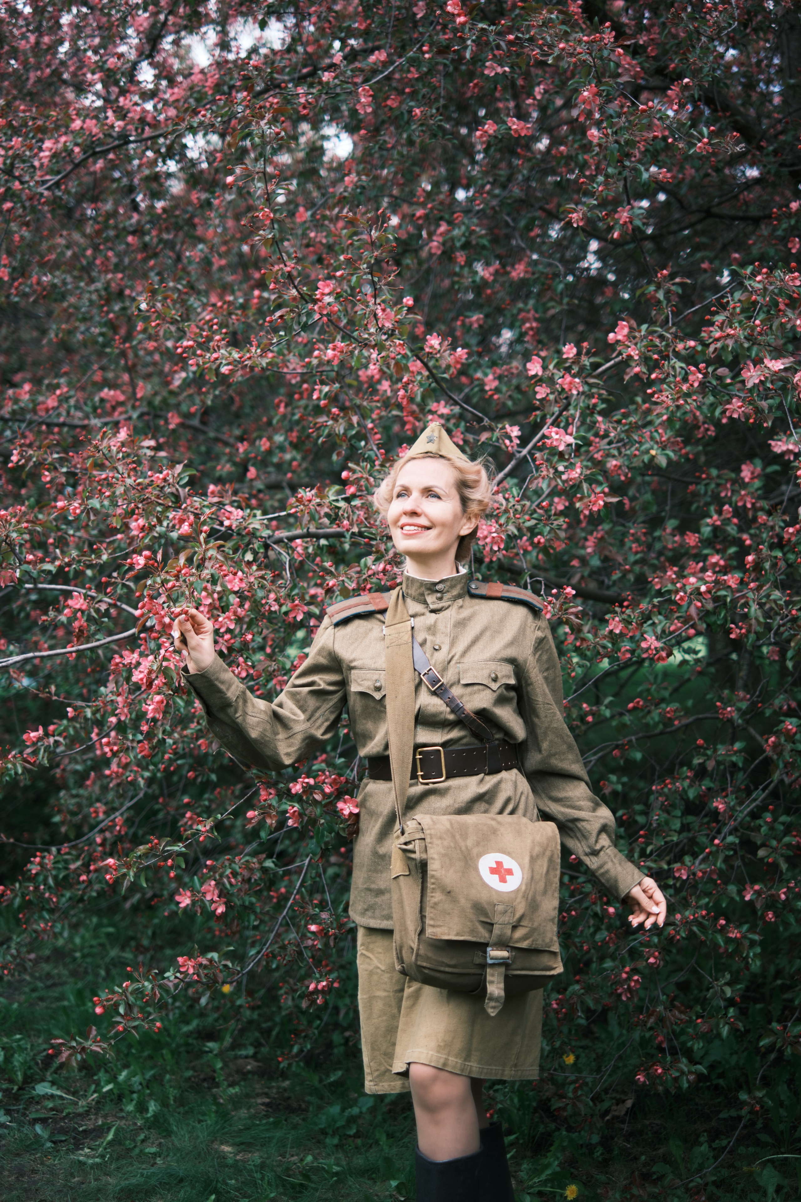 Девушка в военной форме и пилотке стоит на фоне цветущих яблонь