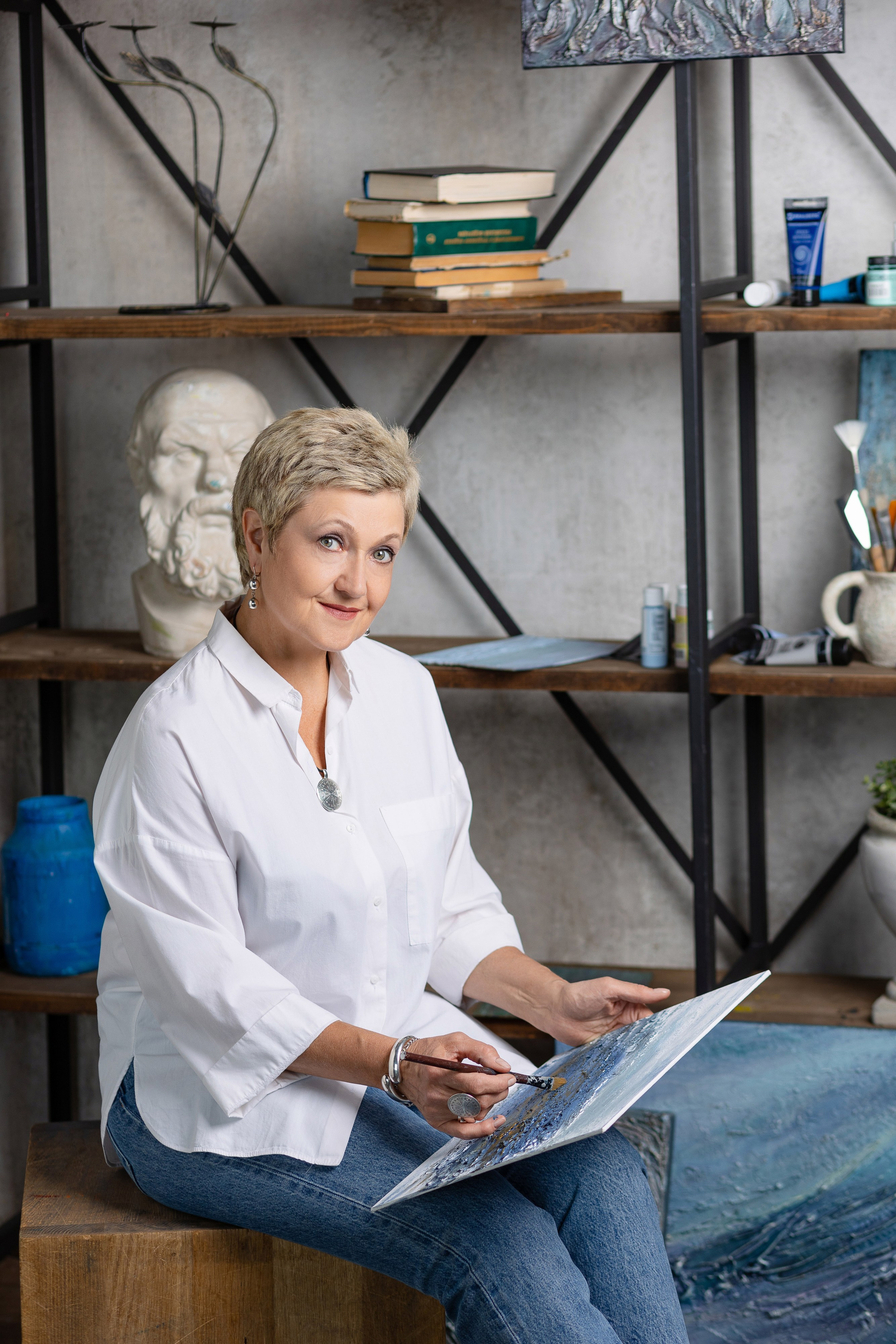 Женщина сидит на диване на фоне стеллажа с книгами и гипсовым бюстом.