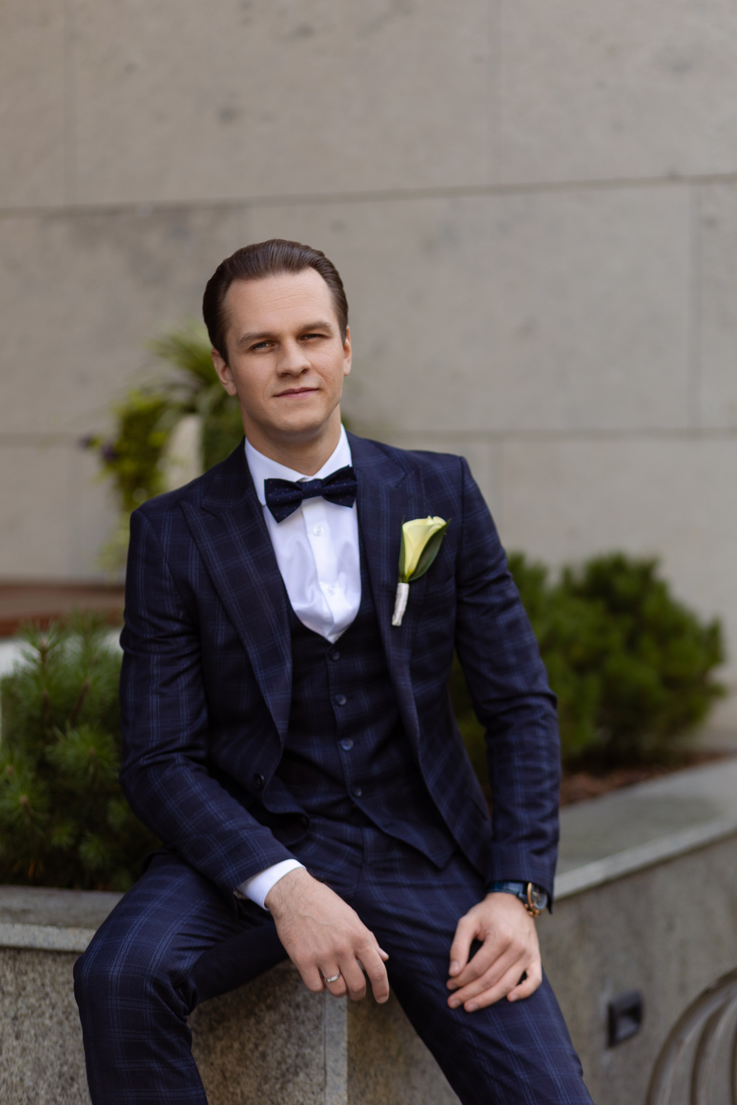 Жених сидит на парапете на фоне зелени.