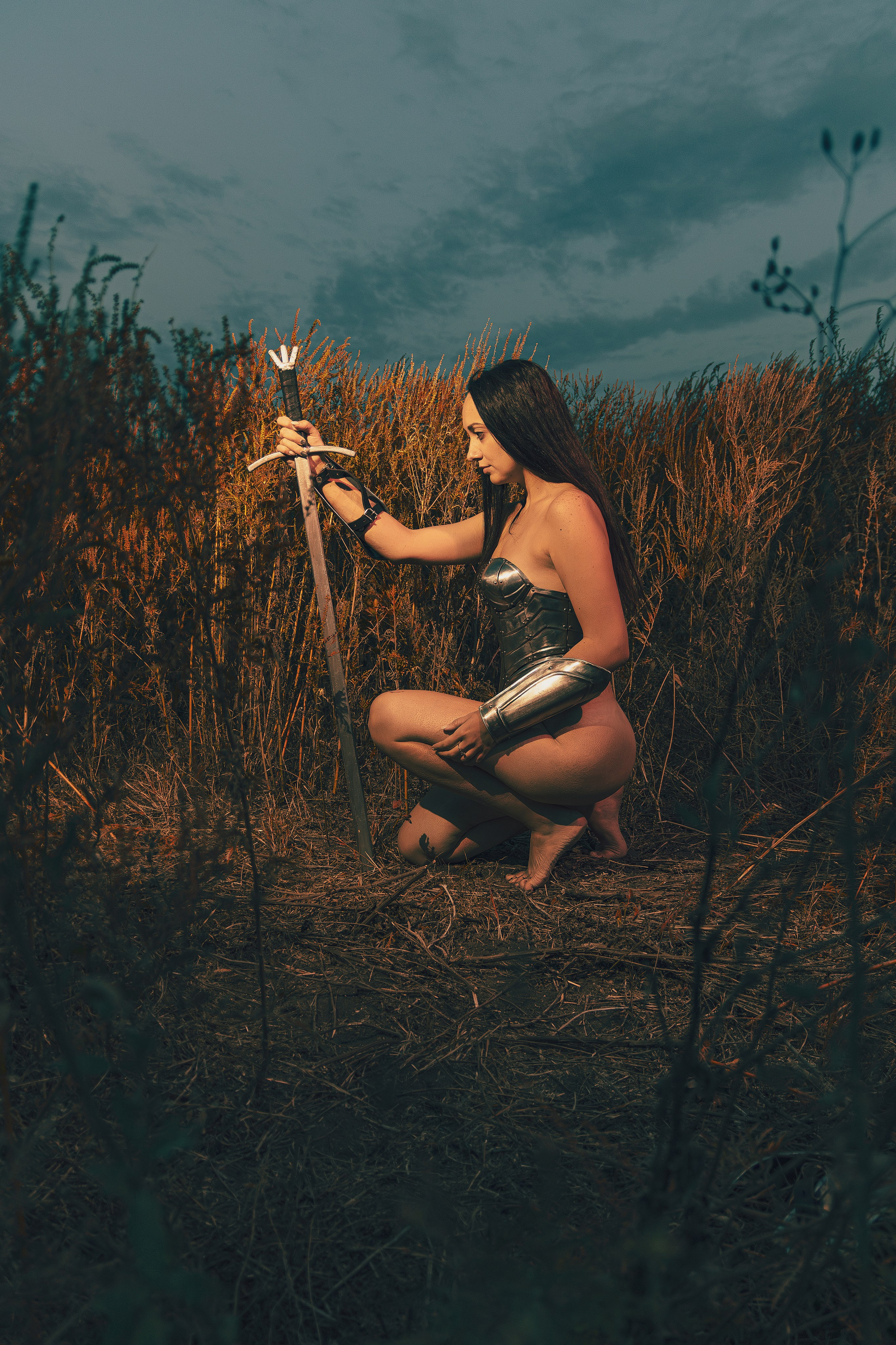 Фотография голой девушки в рыцарских доспехах посреди вечернего неба и высокой жёлтой травы с мечом в руках