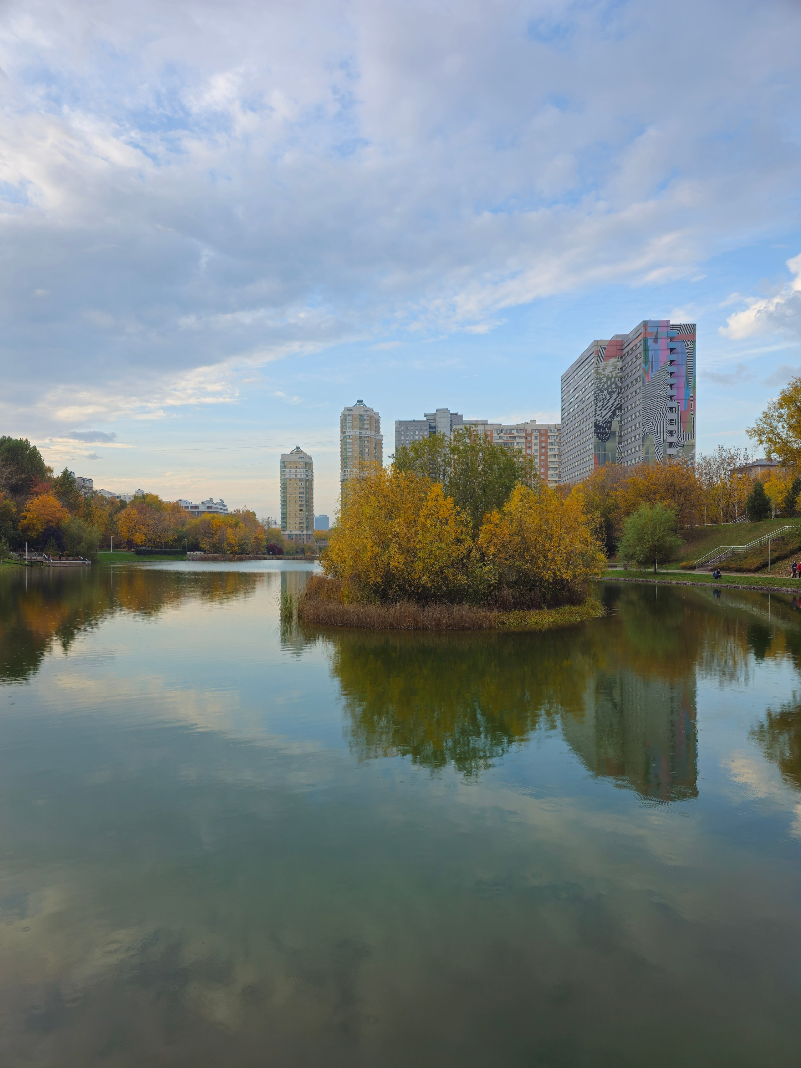 Горизонтальный снимок городского пруда с небольшим островком в центре, покрытым кустами с желтой осенней листвой. На заднем плане, за прудом, видны городские высотки, включая крупное здание с необычным, ярким, многоцветным фасадом (арт-фасад). Вода отражает облачное небо и строения.