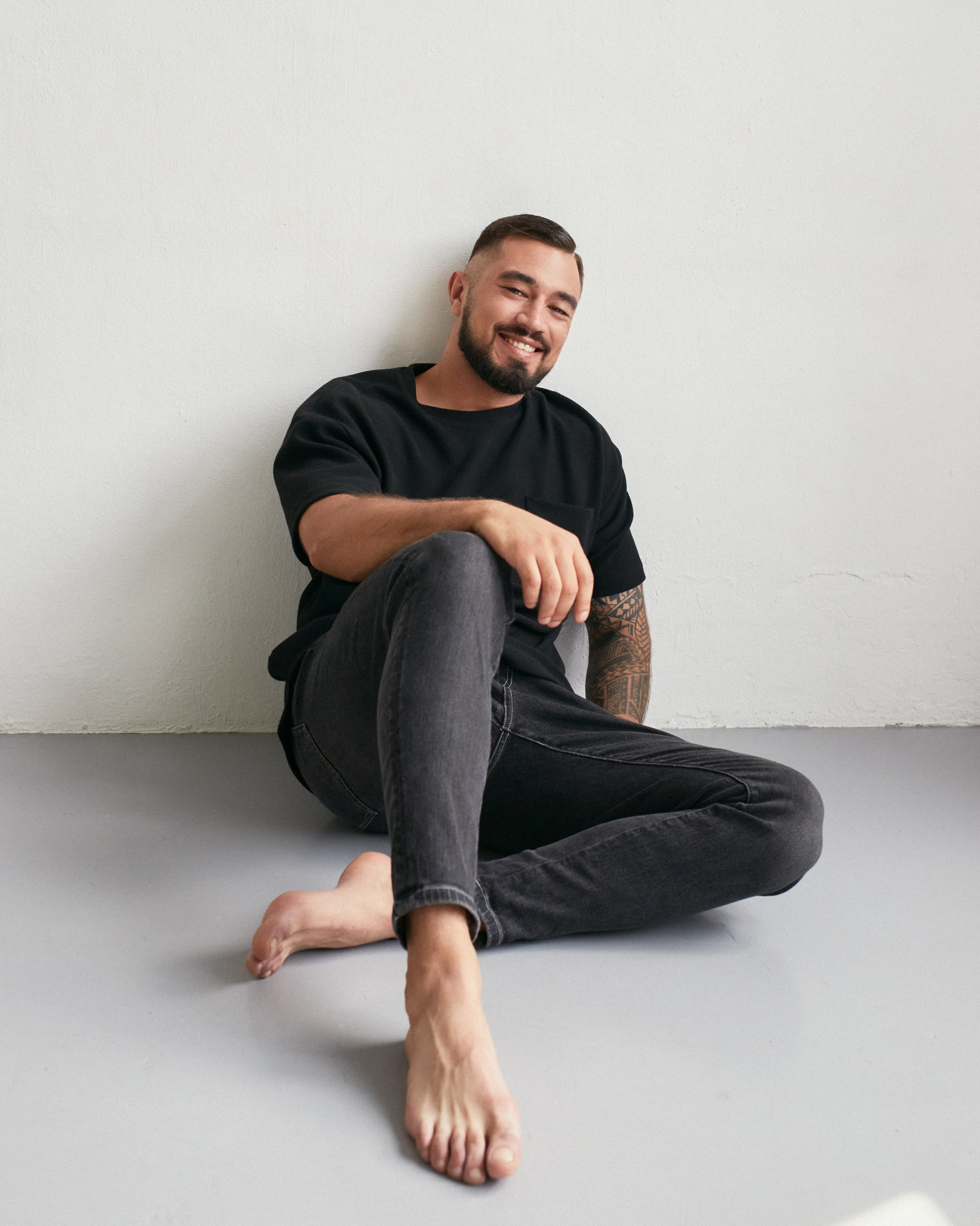 Смуглый мужчина в джинсах и черной футболке сидит на полу и улыбается