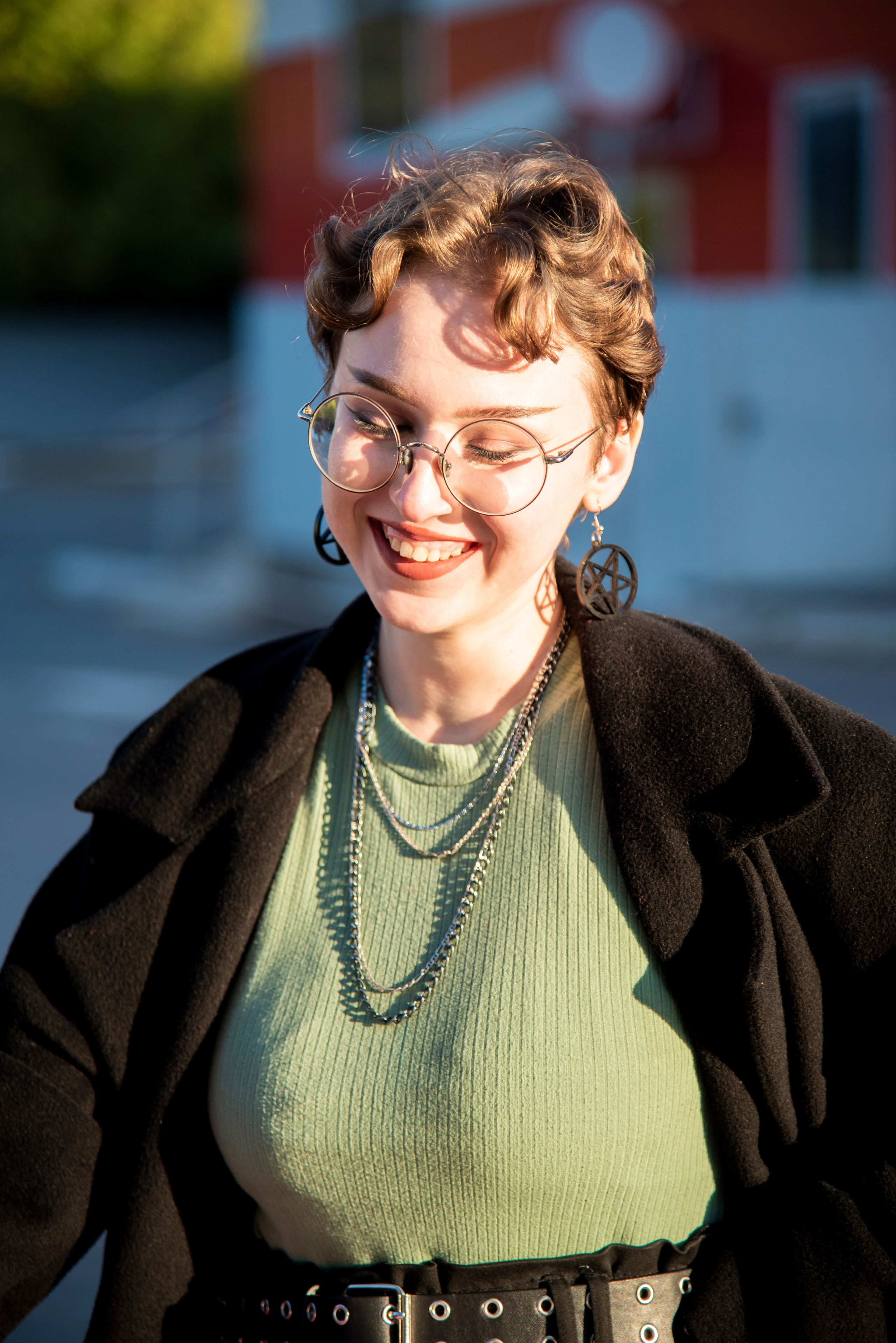 портет девушки. чёрное пальто. красивые позы. серьки в виде пентограммы. короткая стрижка. круглые очки. красный фон