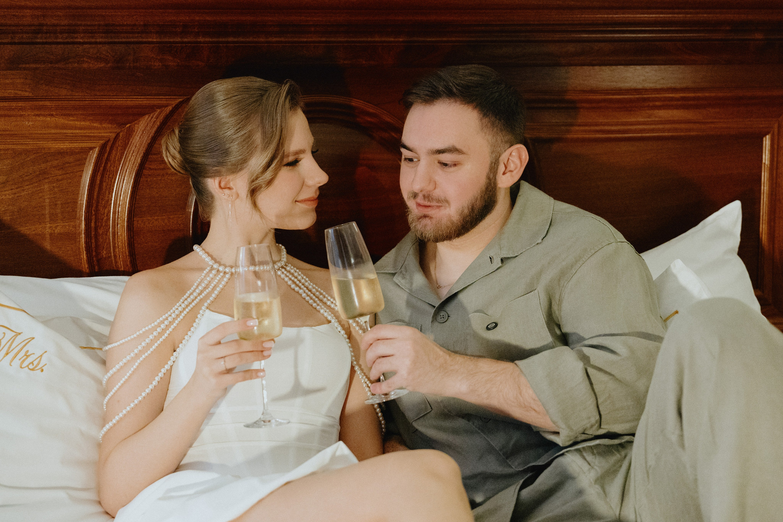 Жених и невеста сидят на кровати, держа бокалы с шампанским.