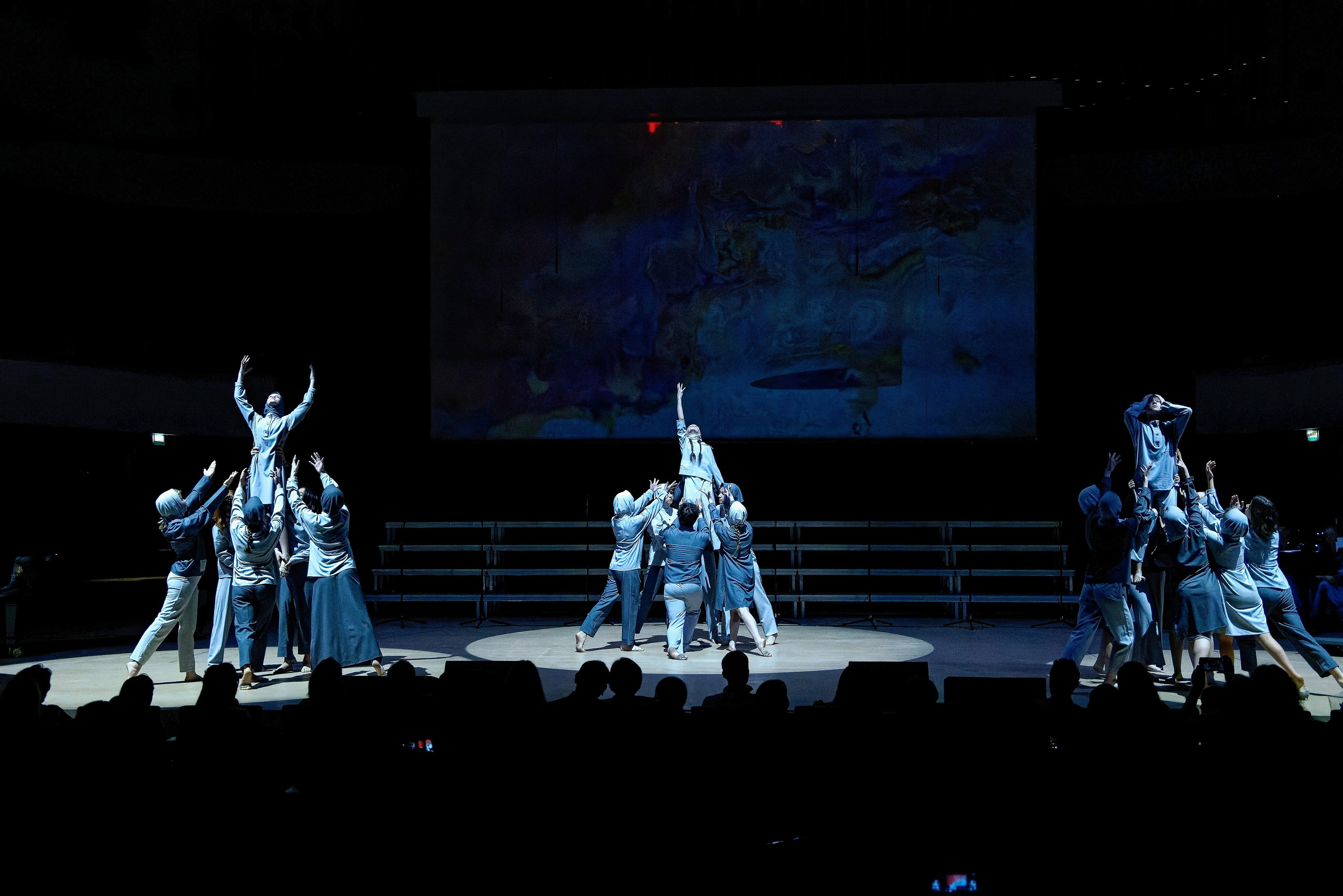 Современный танец контемпорари - группа танцоров в синих костюмах выполняет поддержки и подъемы на сцене с проекцией, художественная хореография