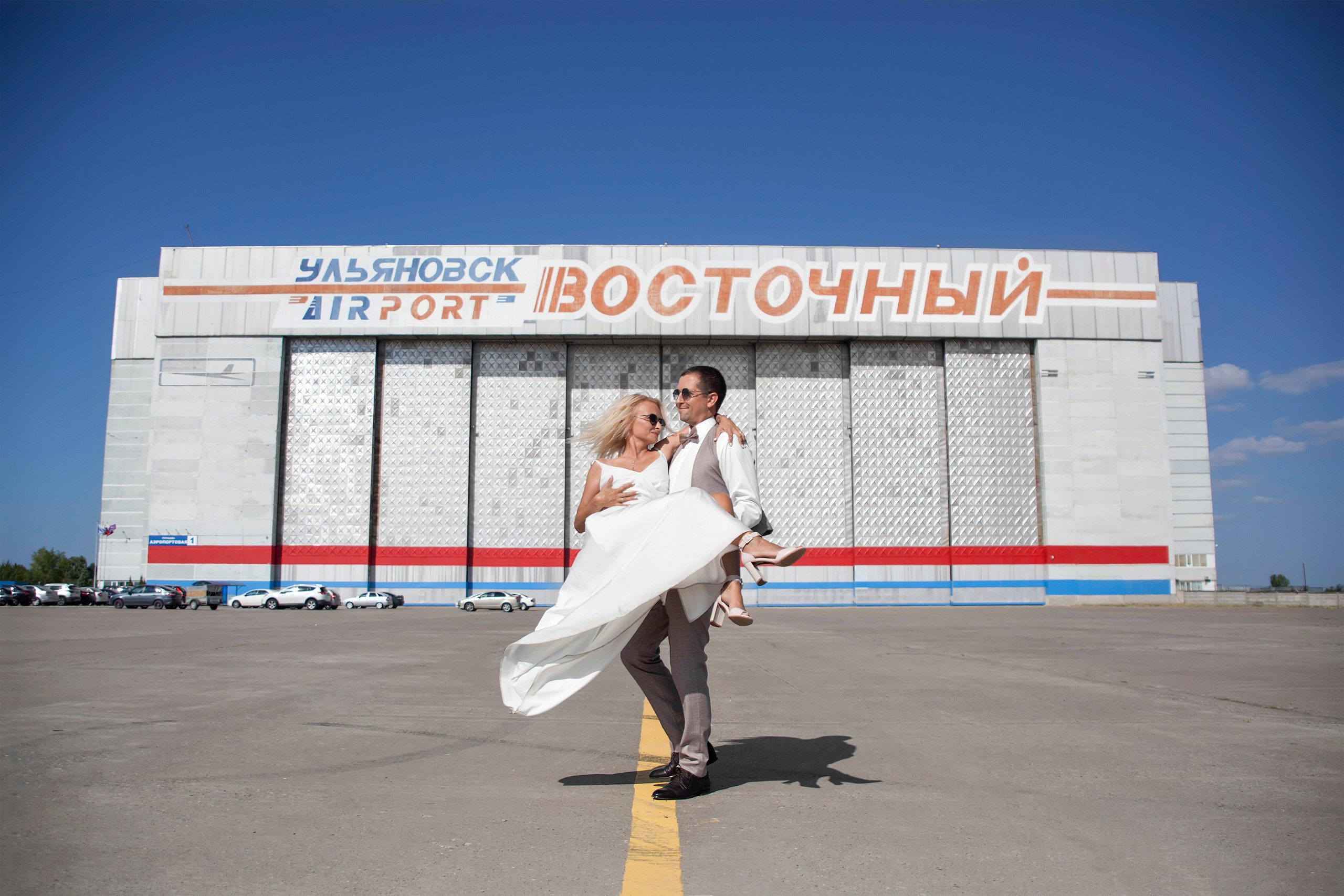 Жених несёт на руках невесту на фоне здания аэропорта, свадебная прогулка Ульяновск