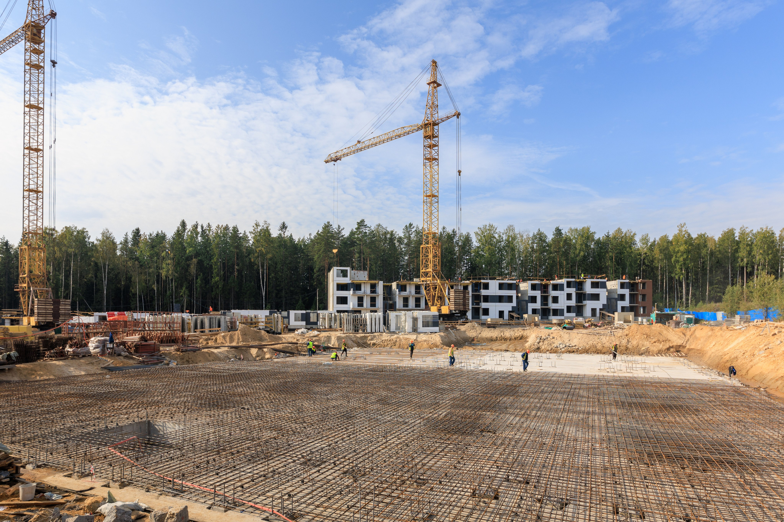 На фотографии изображен строительный объект, окруженный лесом. Видны строительные краны и различные строительные конструкции.