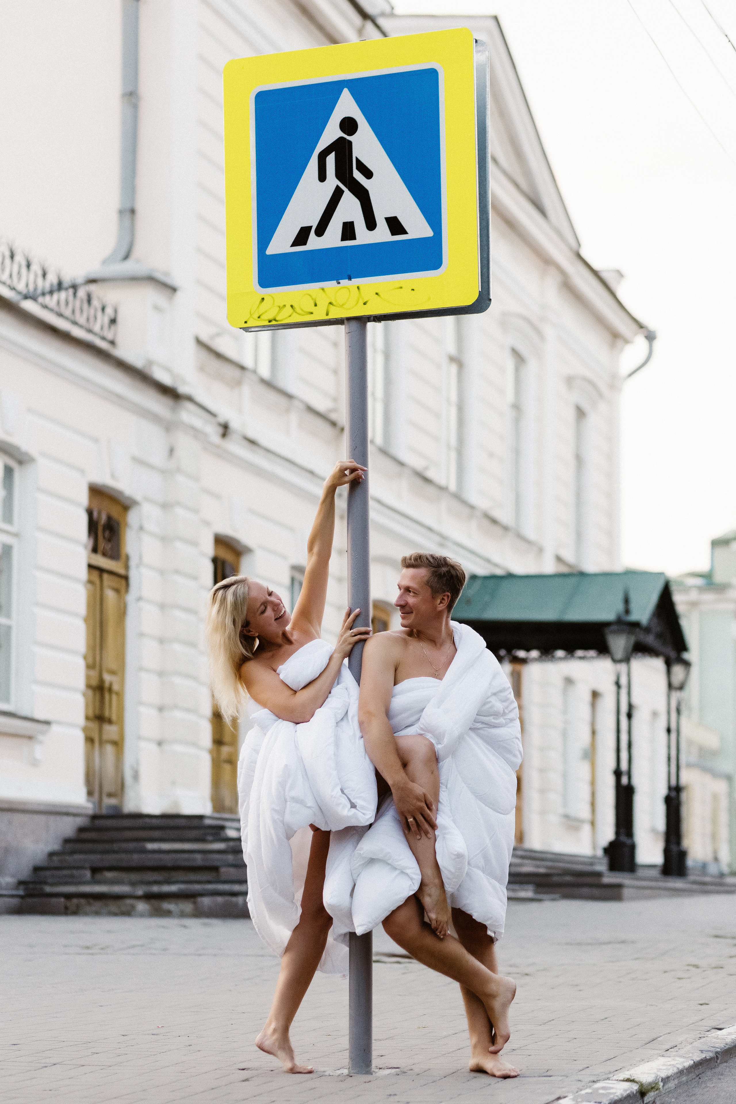 Креативная фотосессия у Театра Чехова в Таганроге: пара в белых одеялах возле дорожного знака на улице Петровская
