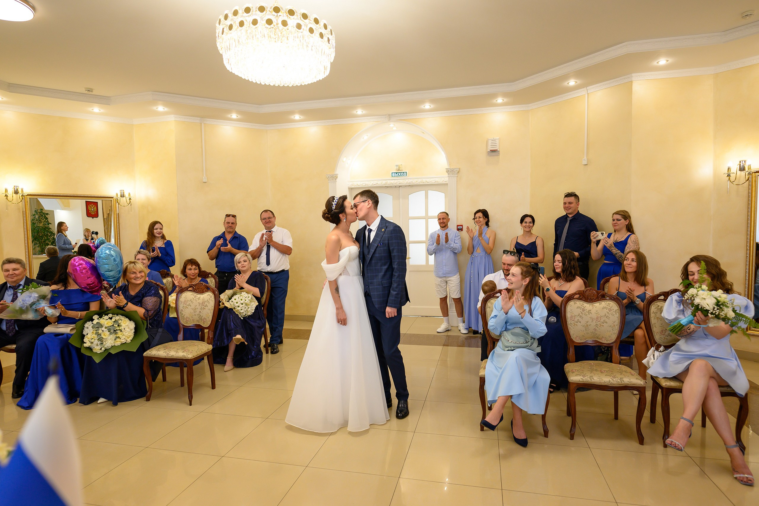 Красивое и эмоциональное фото как молодожены целуются после того как их брак зарегистрировали в ЗАГСе Краснодара, показаны эмоции гостей, кадр сделан на широкий угол.