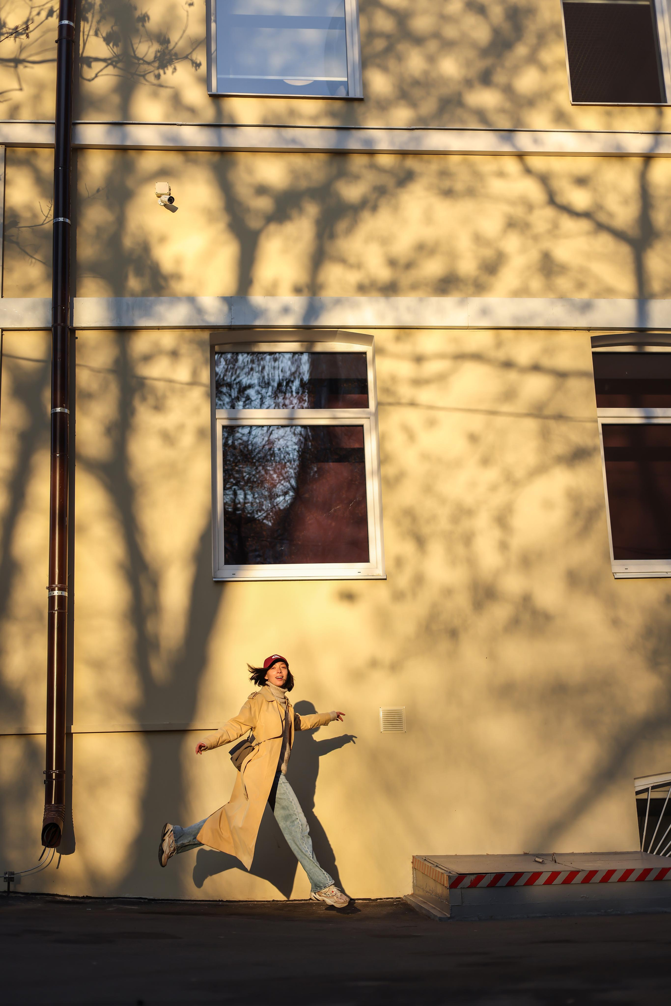 Девушка Наталья Штин на улице в пальто и джинсах в прыжке на фоне дома. В теплом свете