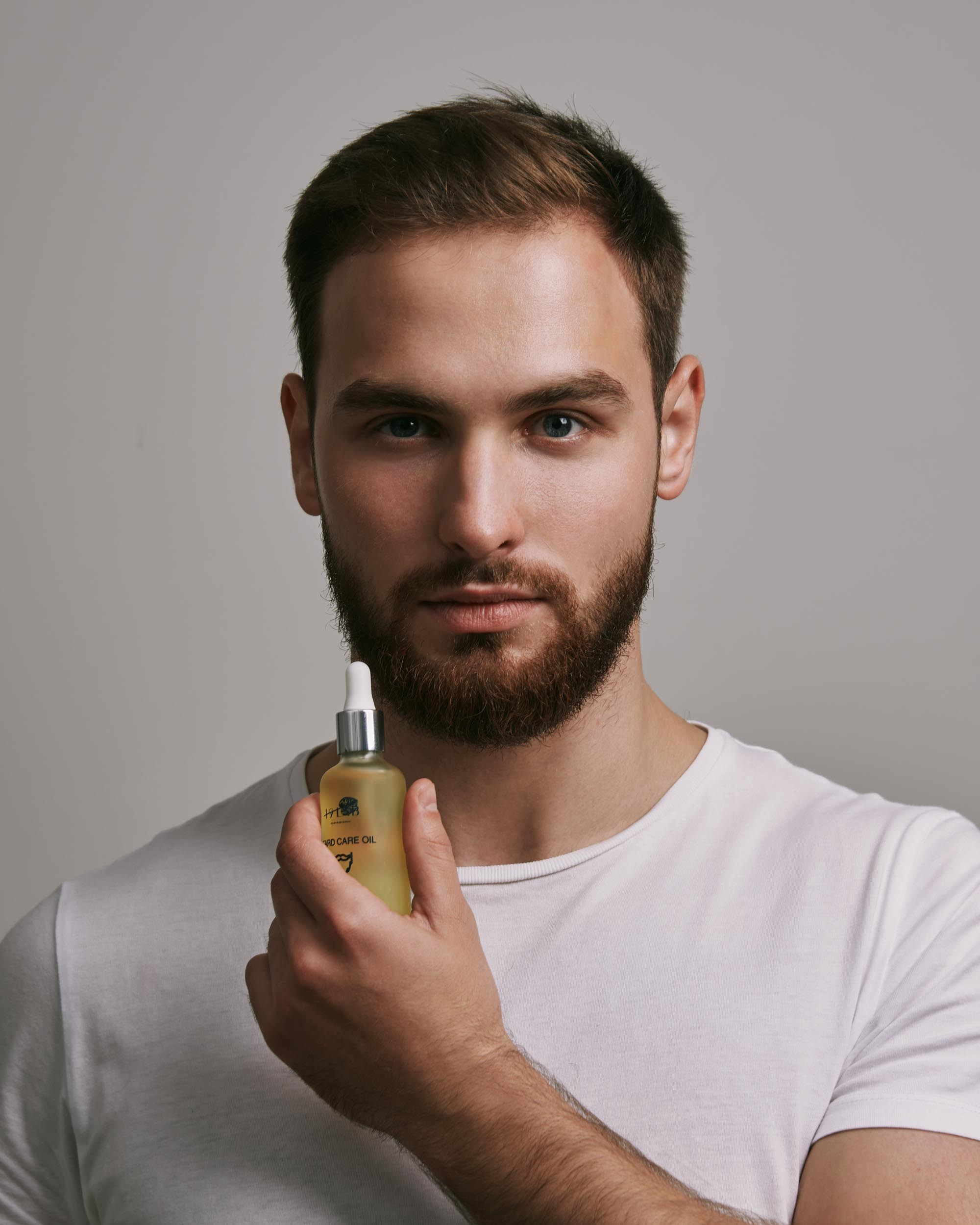 Мужчина с бородой в белой футболке держит в руках флакон с косметикой