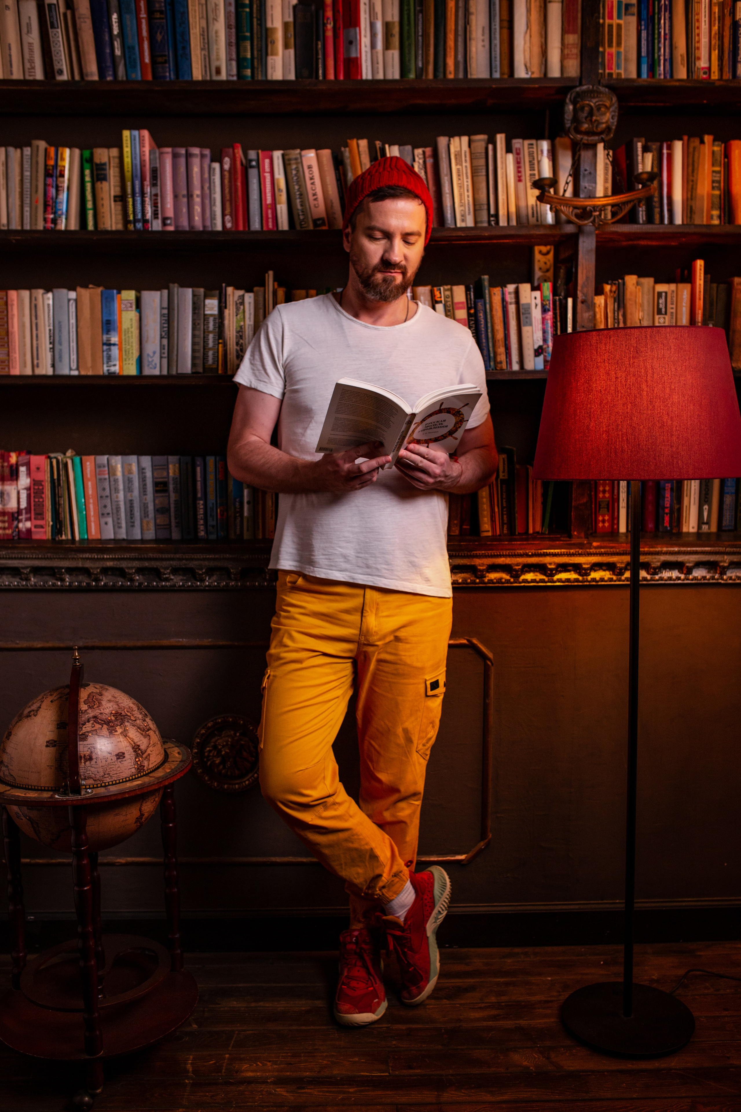 Парень в яркой одежде и в красной шапке бини читает книгу в библиотеке.